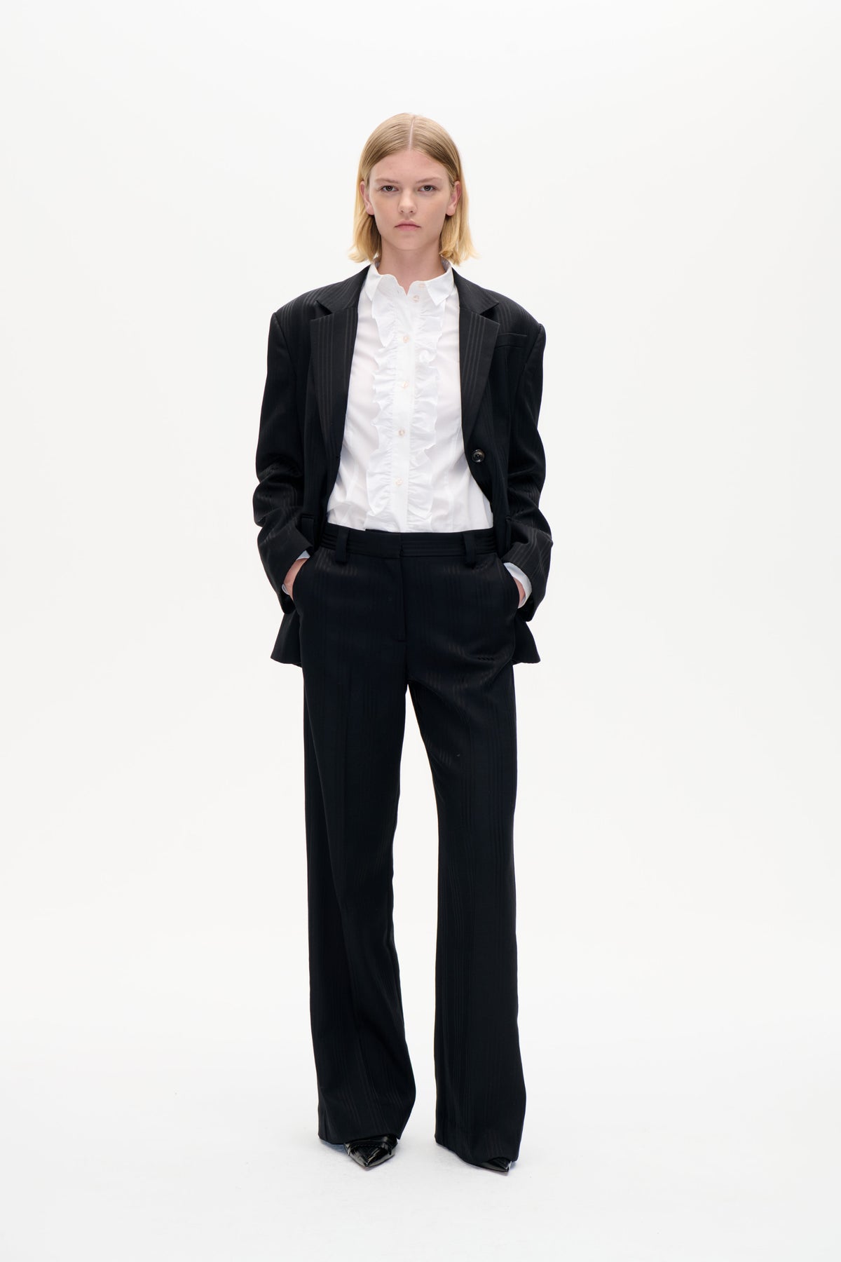 Front-facing model in a black pinstripe blazer and wide-leg trousers over a white ruffled button-up shirt, against a plain white background.