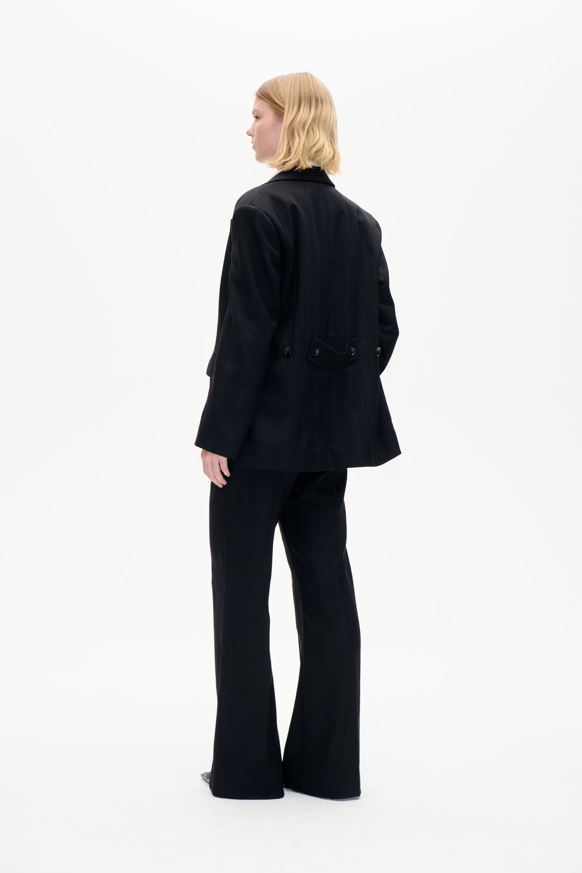 Model in a black pinstripe blazer with a buttoned back waist tab and wide-leg trousers, shown from the side and back.