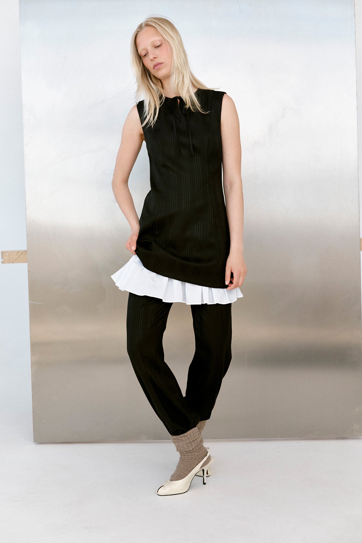 Model wearing a sleeveless black tunic with subtle vertical stripes and a tie neckline over a white ruffled hem and matching trousers.
