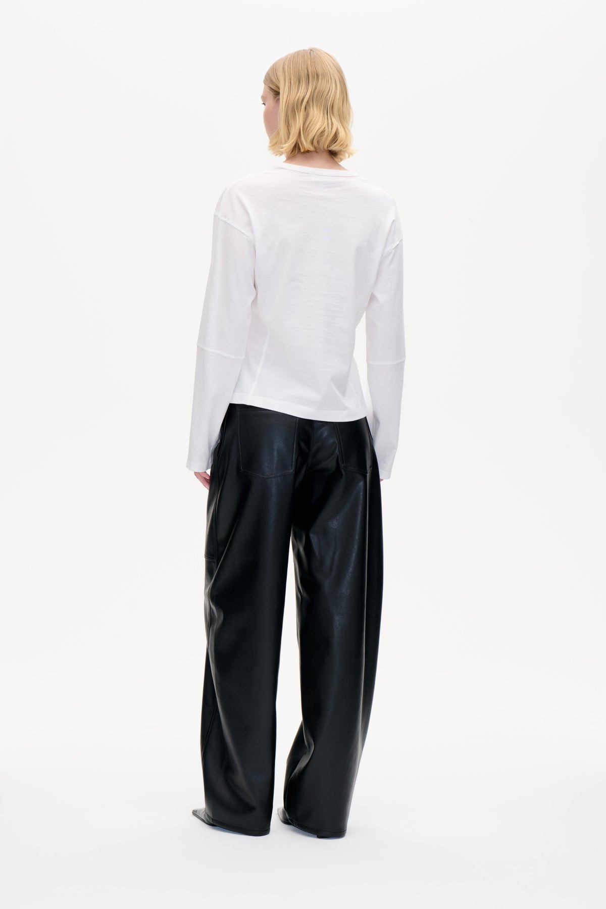 Back view of a model wearing a plain white long-sleeved top and glossy black wide-leg trousers with patch pockets.