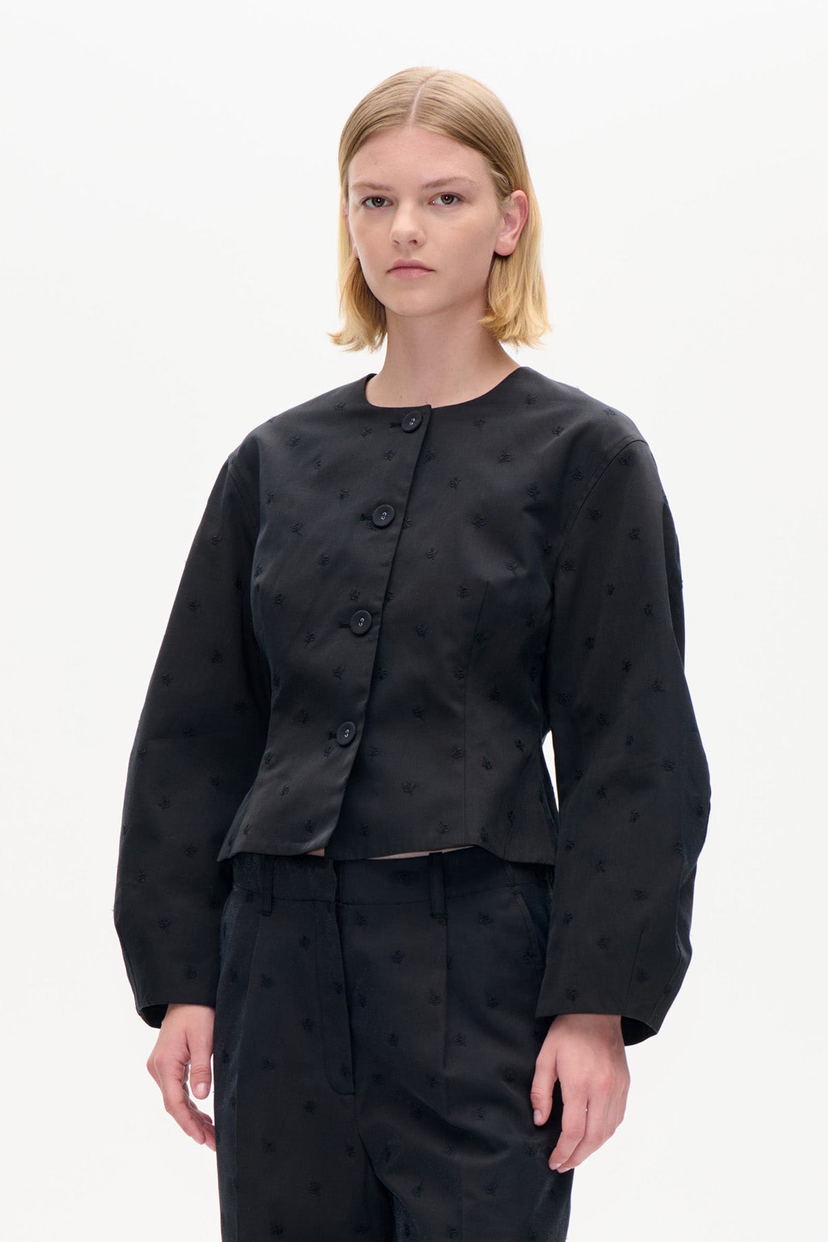 Model wearing a black cropped jacket with subtle embroidered motifs, a round neckline and large buttons, paired with matching trousers.