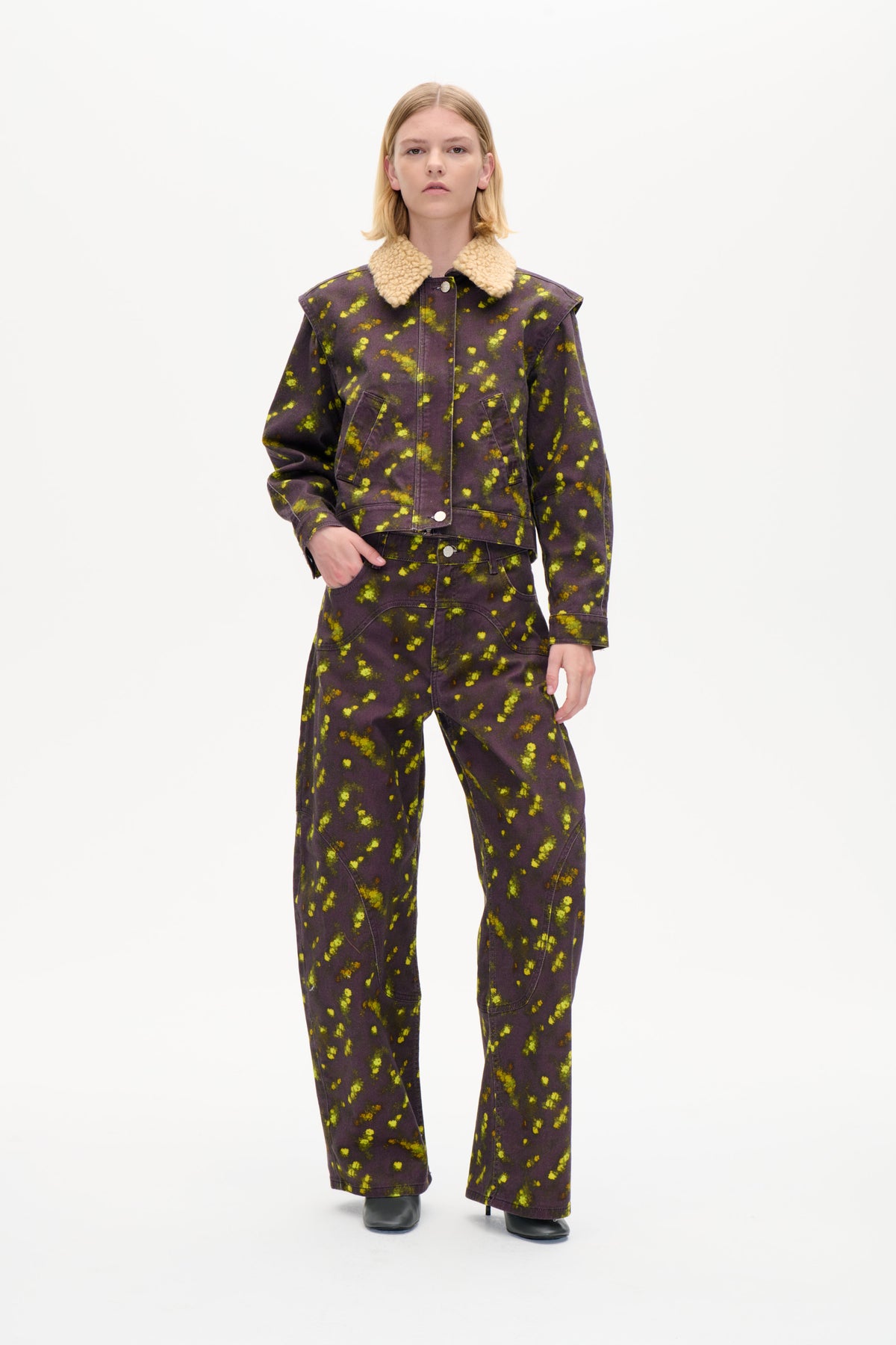 Front view of a model wearing a charcoal-purple jacket with bright yellow mottled print and a cream textured collar, with matching wide-leg trousers.