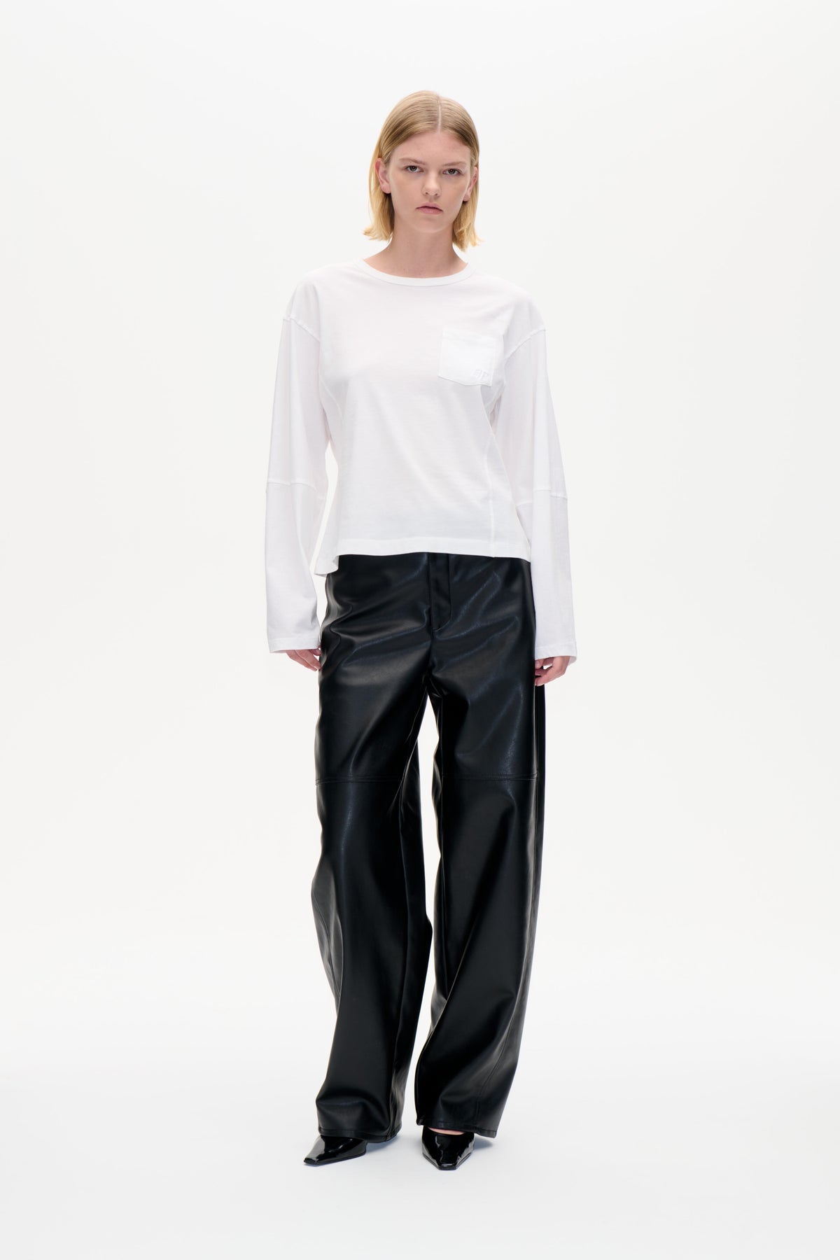 Front-facing model in a white long-sleeved T-shirt with chest pocket and black wide-leg trousers against a plain white backdrop.