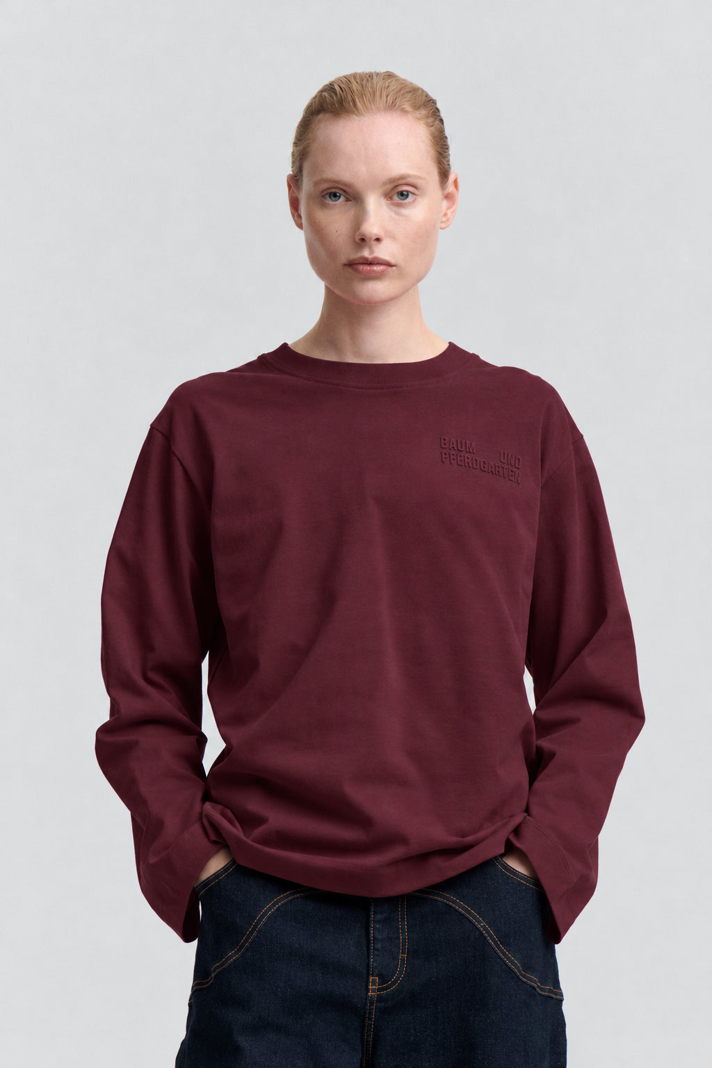 Front view of a burgundy long-sleeved crew-neck sweatshirt with subtle tonal chest lettering, styled with dark jeans.