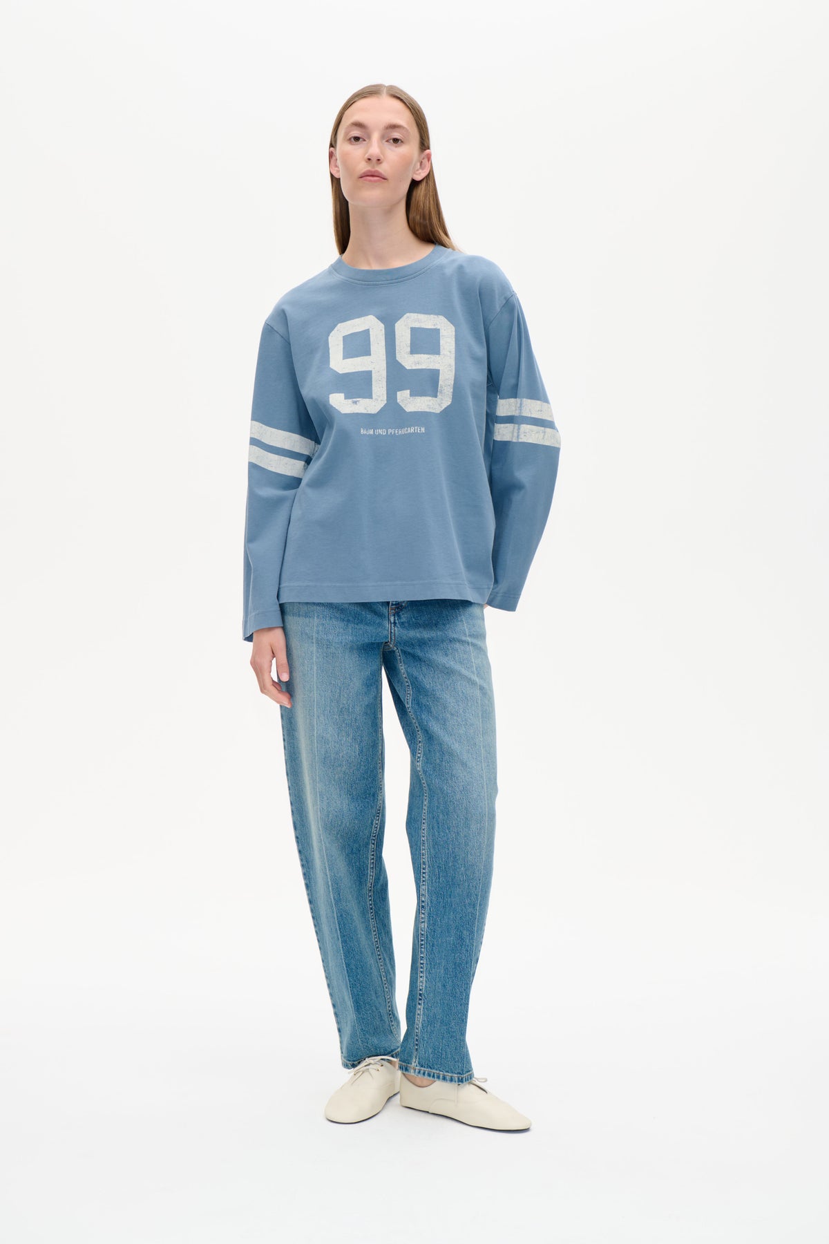 Front view of a model wearing a loose blue sweatshirt with a large distressed white 99 graphic and striped sleeves, paired with jeans.
