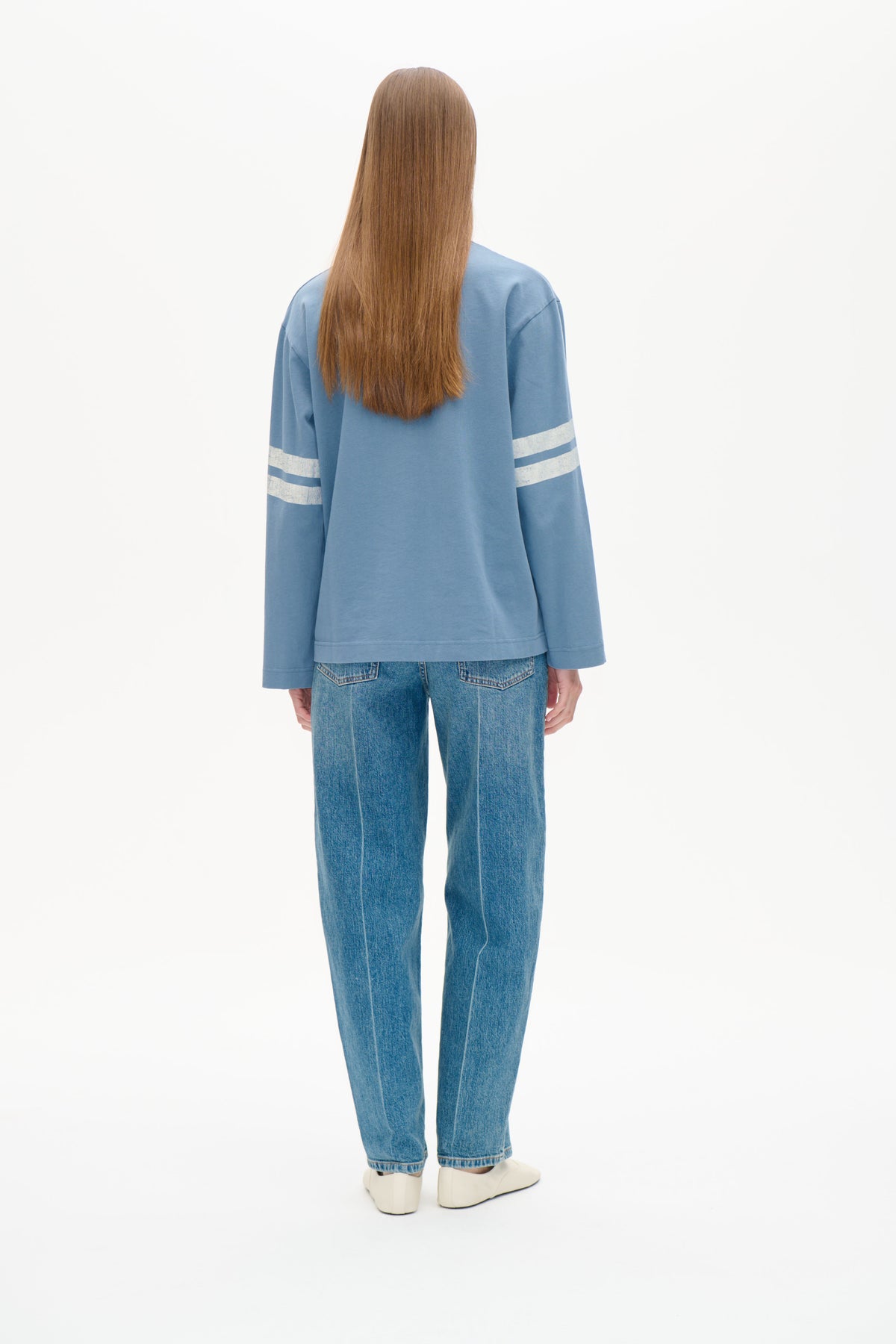 Back view of a model wearing a light blue long-sleeve sweatshirt with white sleeve stripes and blue jeans.
