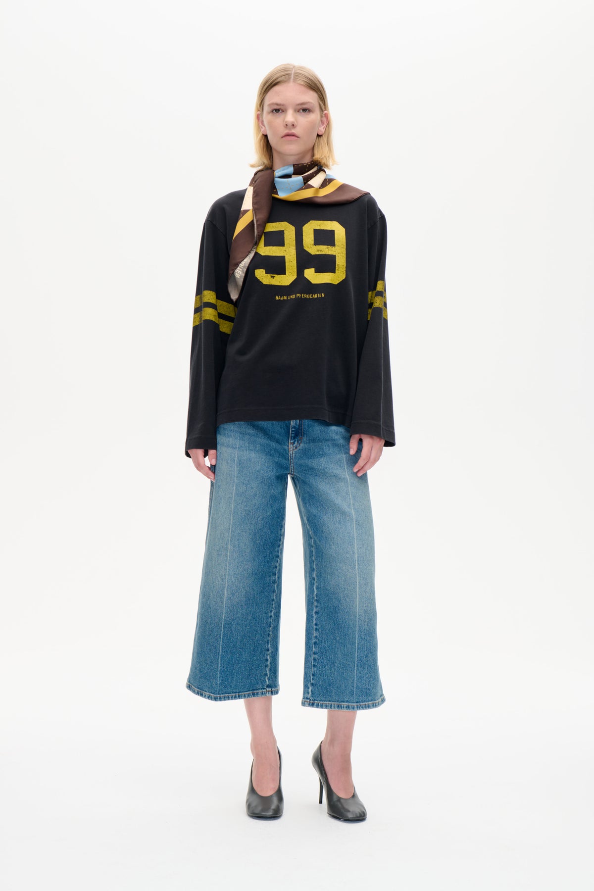 Model wearing a charcoal long-sleeved sweatshirt with a large yellow 99 graphic, wide-leg blue jeans and a multicoloured geometric scarf.