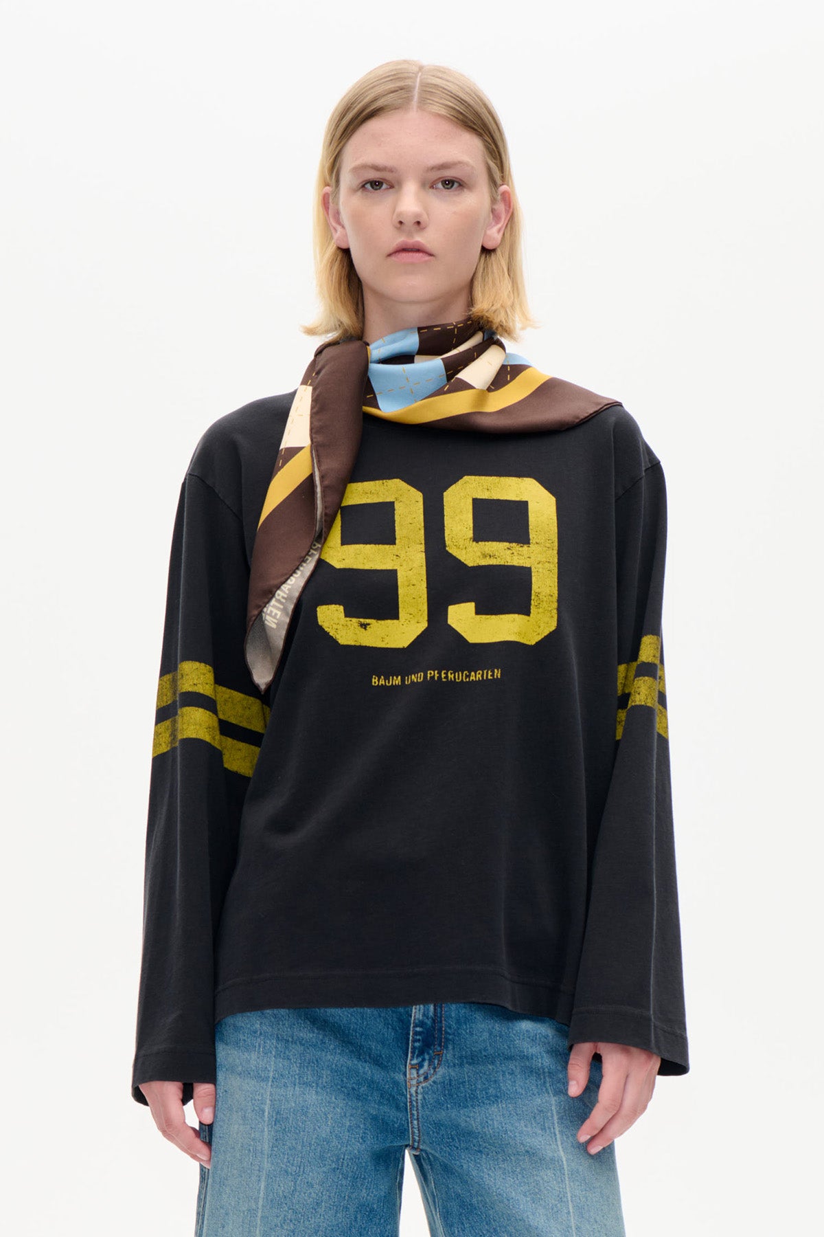 Front view of a model wearing a black long-sleeve top with yellow number graphics, a multicoloured patterned scarf and blue jeans.