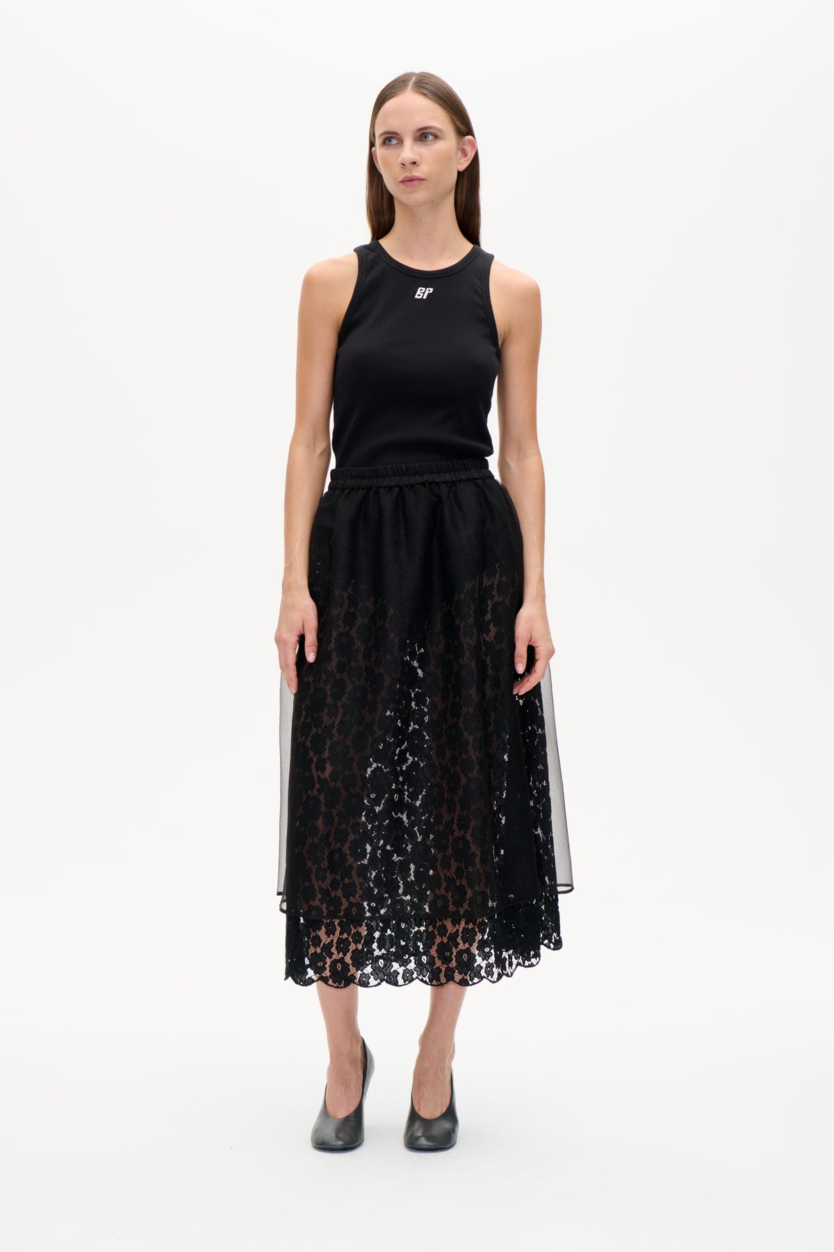 Front view of a model wearing a black ribbed sleeveless tank and a black floral lace skirt with a sheer overlay and scalloped hem.