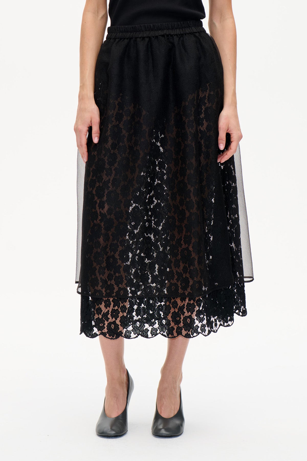 Front view of a black midi skirt with floral lace, a scalloped hem and a sheer overlay, worn with a black top.