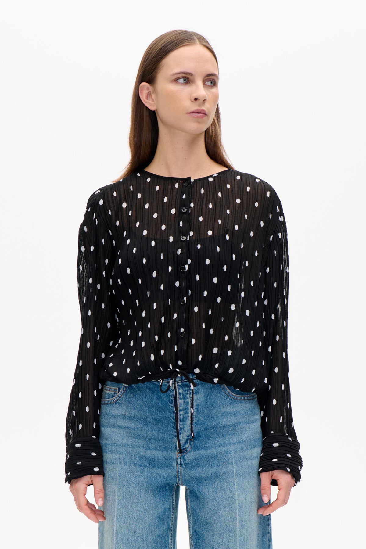 Front view of a black sheer, crinkled blouse with white irregular dots, a button front and long sleeves, worn with blue jeans.