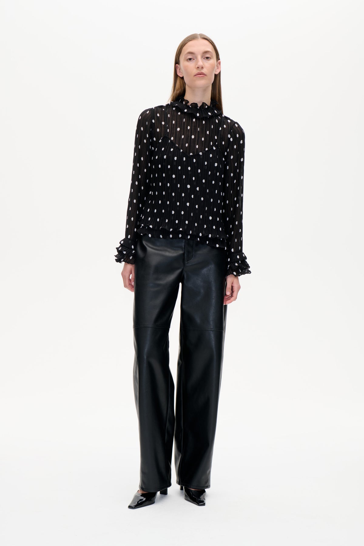 Front-facing model in a sheer black blouse with white polka dots and ruffled trim, paired with black wide-leg trousers.