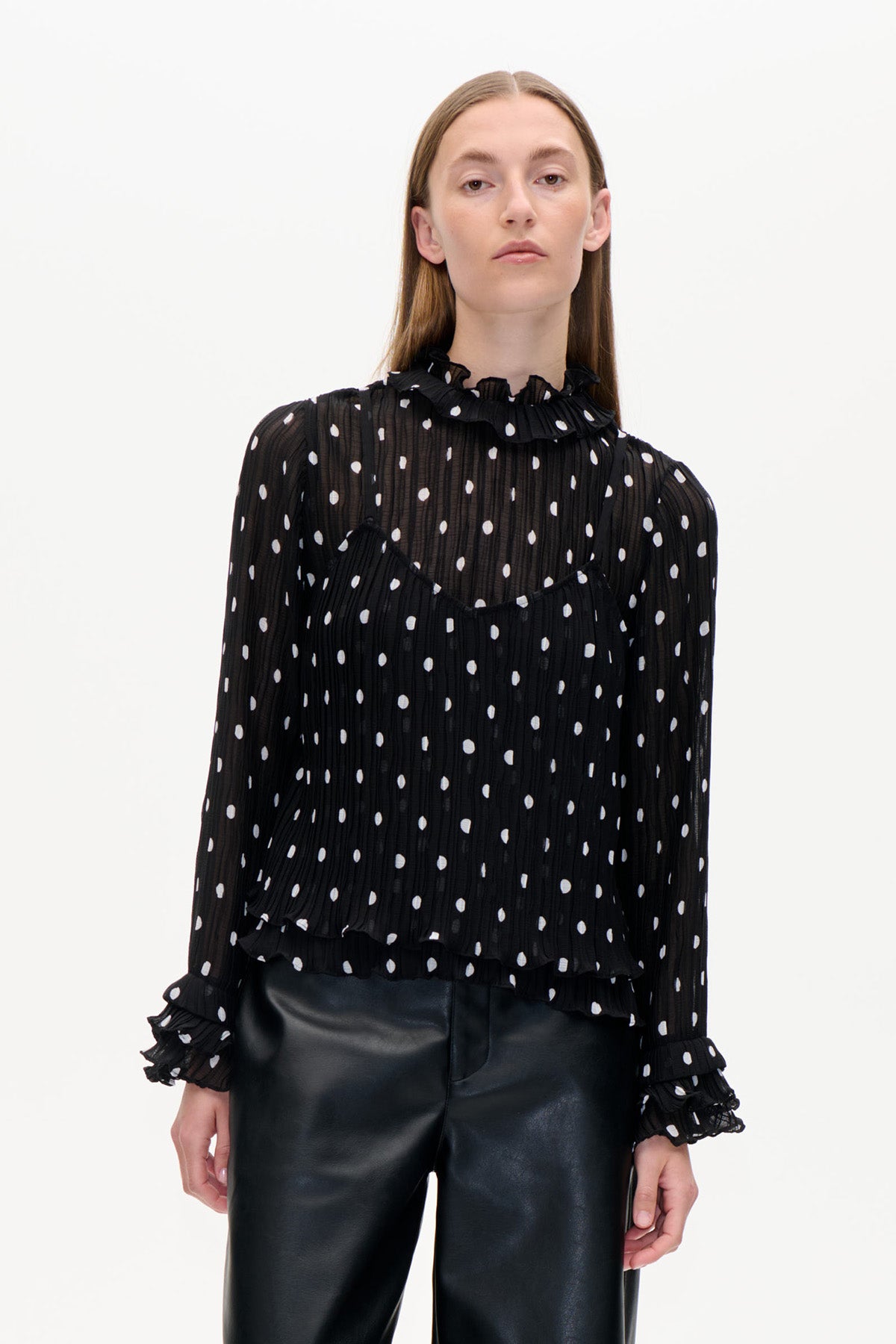 Front view of a black sheer, textured blouse with white polka dots, a ruffled high neck and cuffs, paired with black trousers.