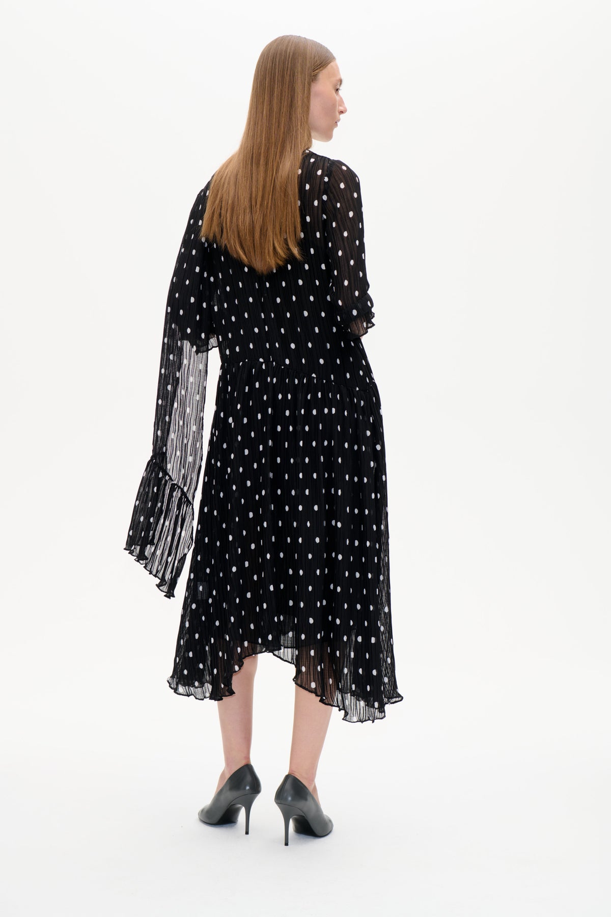 Model in a black sheer pleated midi dress with white dots and ruffled sleeves, shown from the back in black heels.