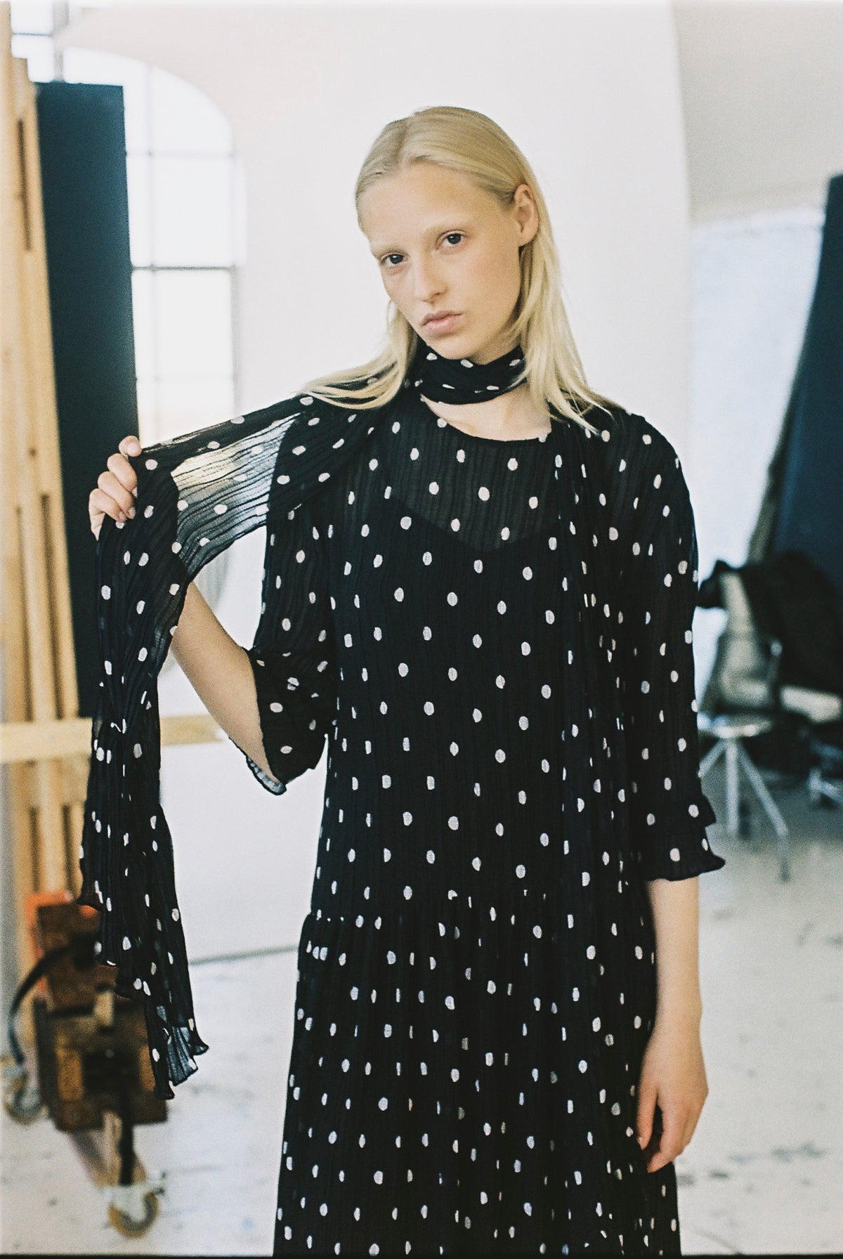 Model wearing a black dress with irregular white dots, a high neckline and sheer pleated sleeves, holding one sleeve out.