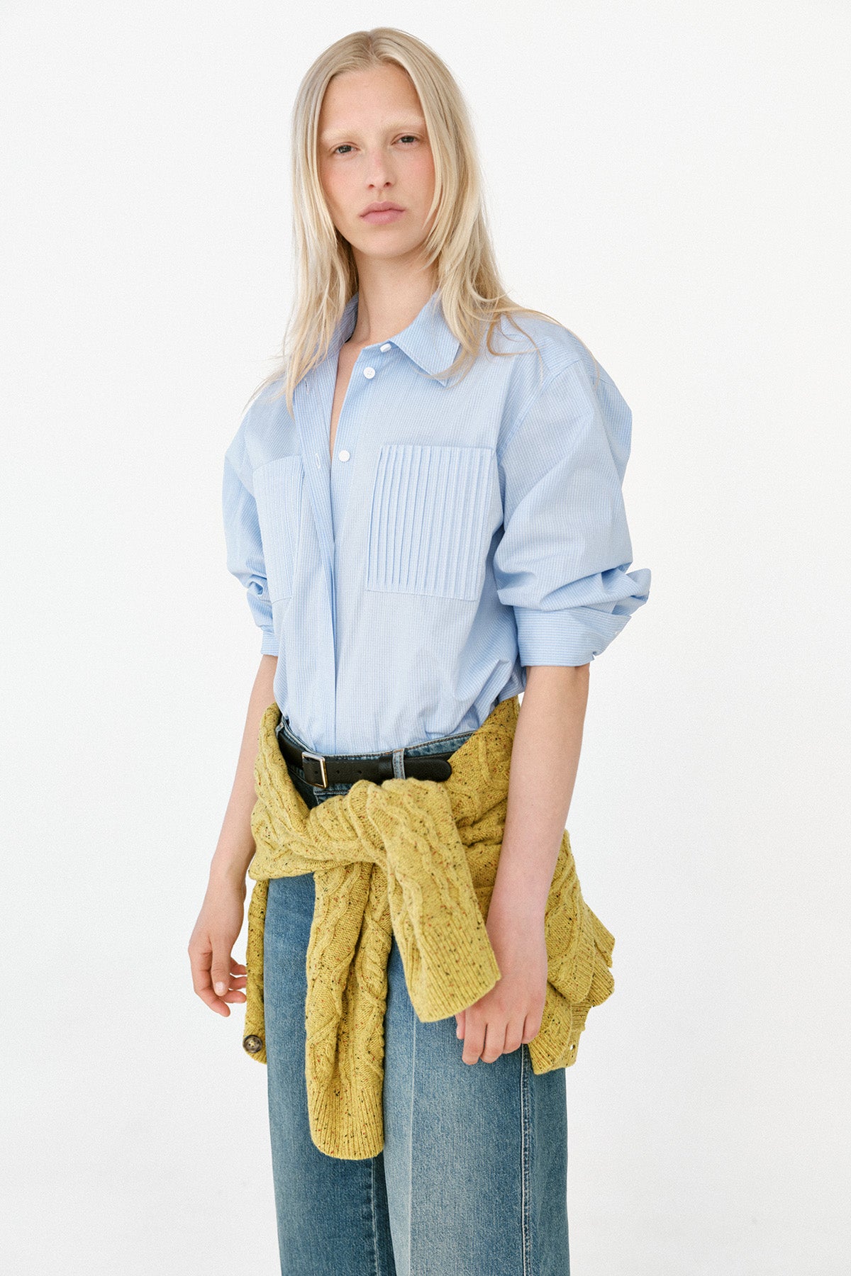 Model wearing a pale blue oversized button-up shirt with a pleated chest pocket, wide-leg blue jeans and a mustard knit tied at the waist.
