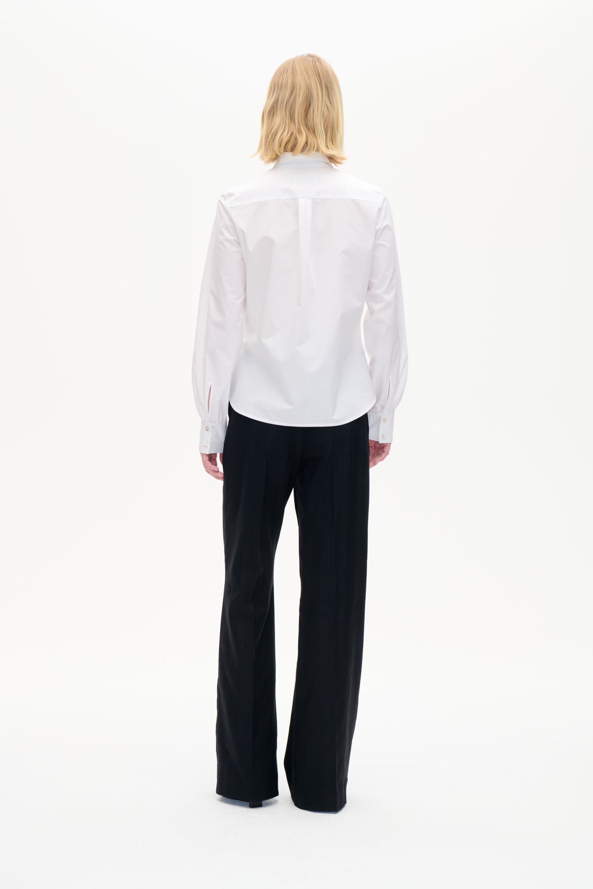 Back view of a white long-sleeved button-up shirt with a back yoke and buttoned cuffs, paired with black trousers.