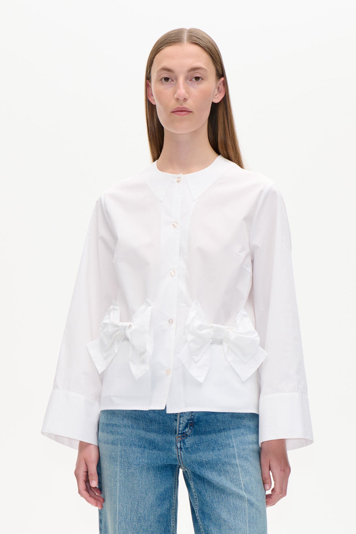 Vorderansicht einer weißen Bluse mit durchgehender Knopfleiste, spitzem Kragen, oversized Manschetten und großen Schleifen in der Taille, kombiniert mit blauen Jeans.