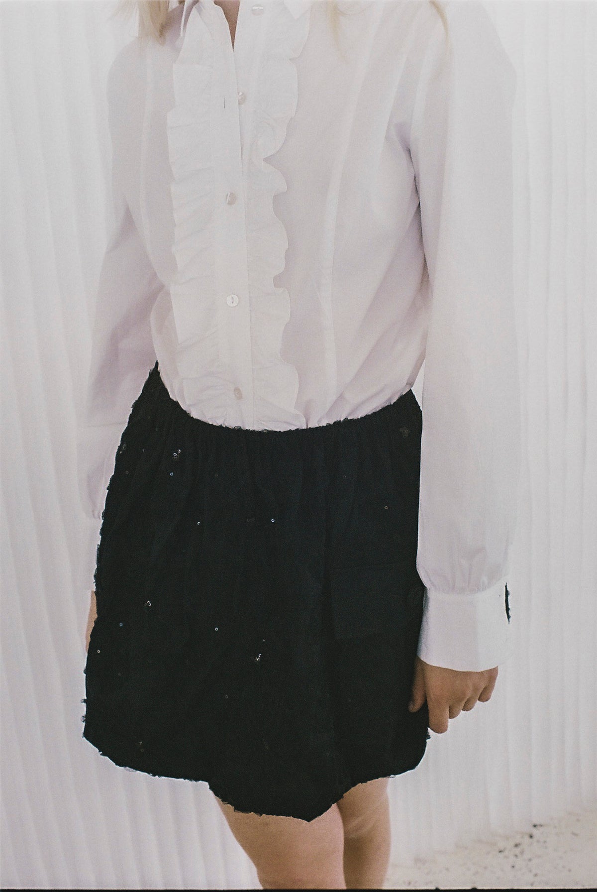 Cropped campaign view of a model wearing a white ruffled button-front blouse and a black skirt with scattered sequins.