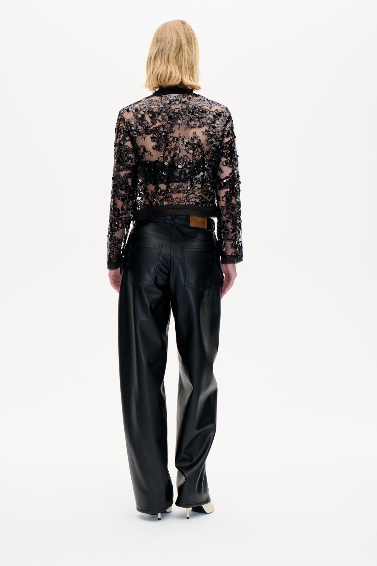 Back view of a model wearing a sheer black long-sleeved top covered in sequins and wide-leg black trousers.