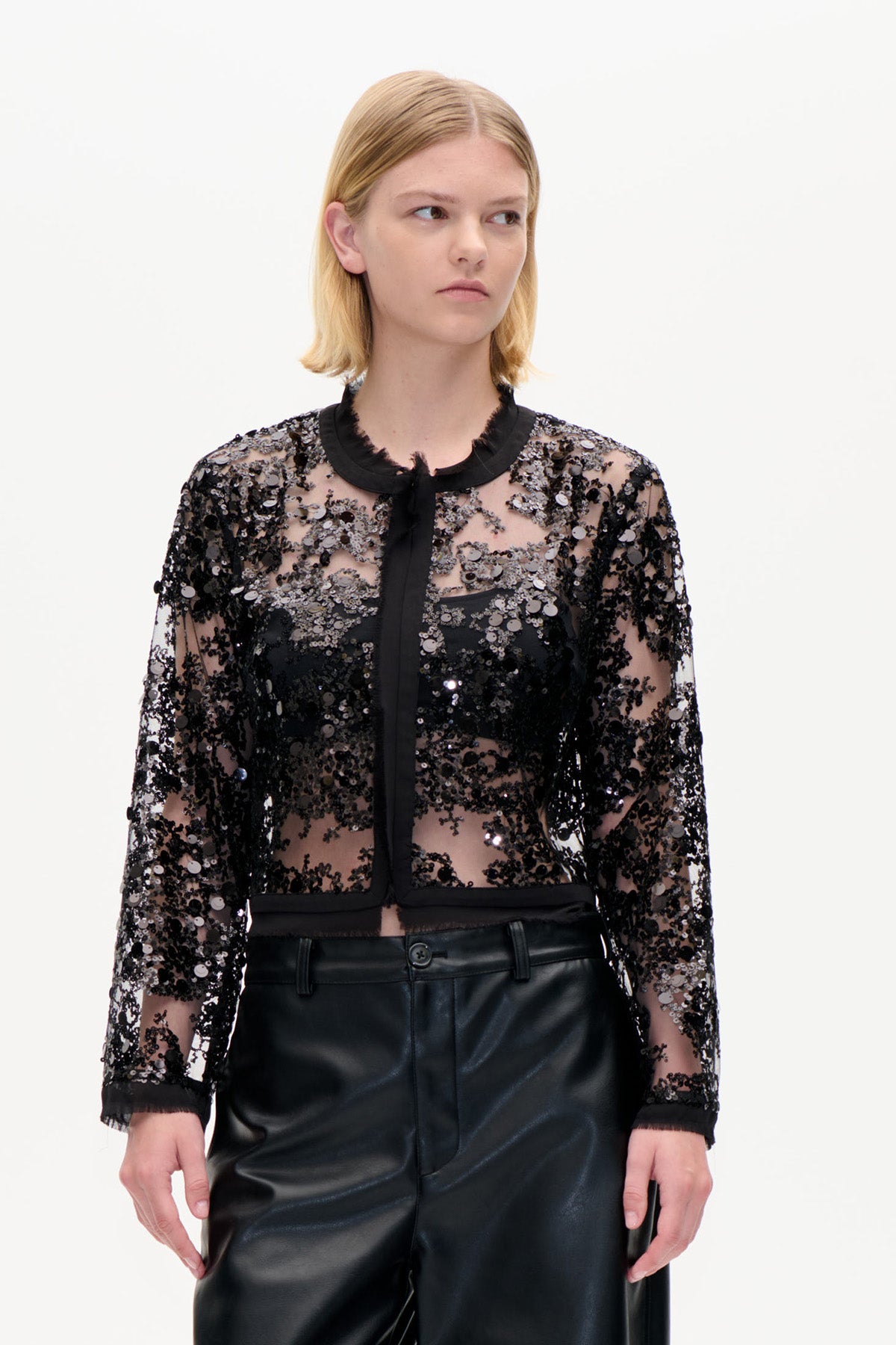 Front view of a sheer black long-sleeved blouse covered in sequins, with a black trim collar and button-free front, paired with black trousers.