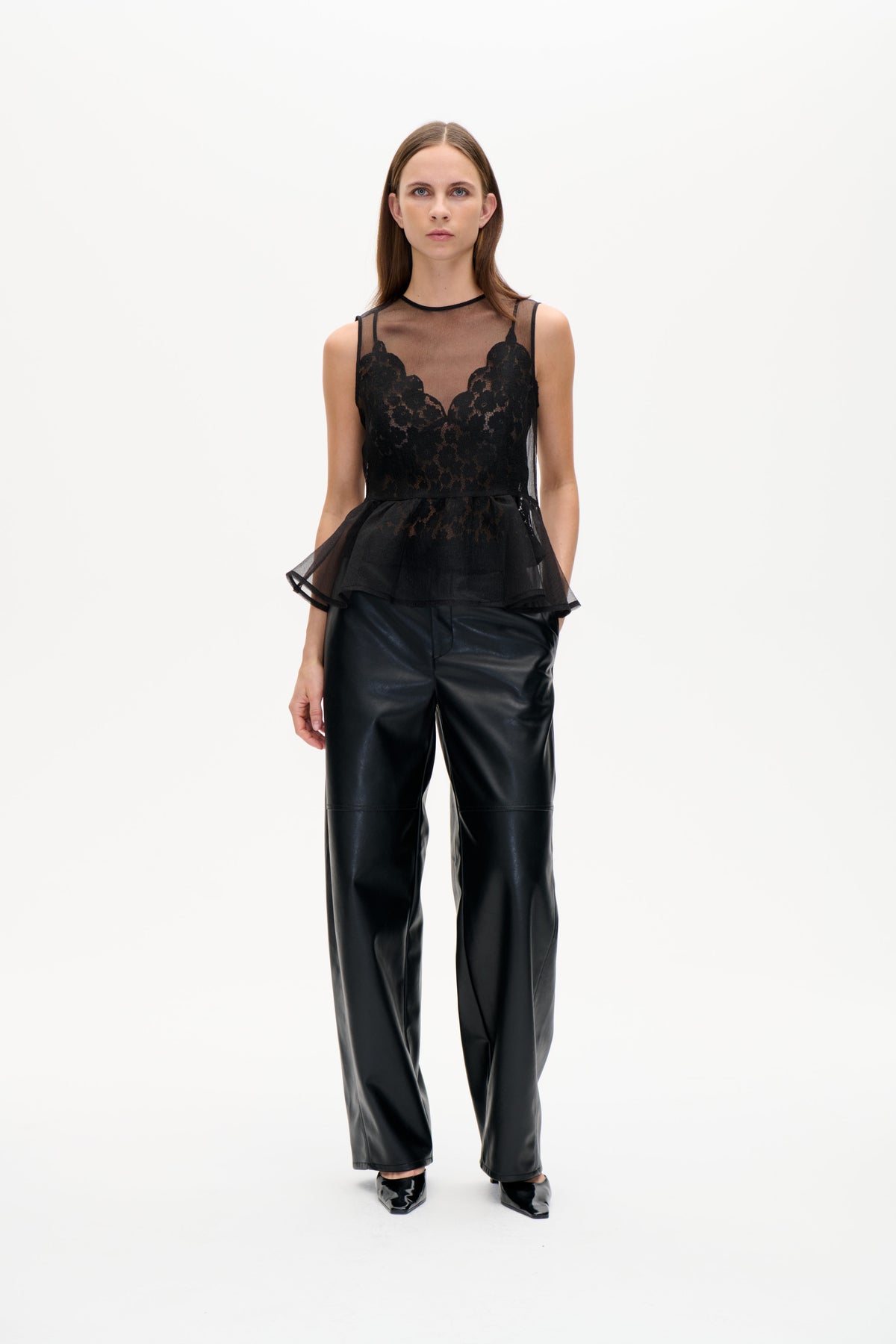 Model wearing a black sheer sleeveless top with floral lace panels and a flared peplum, paired with glossy wide-leg trousers.