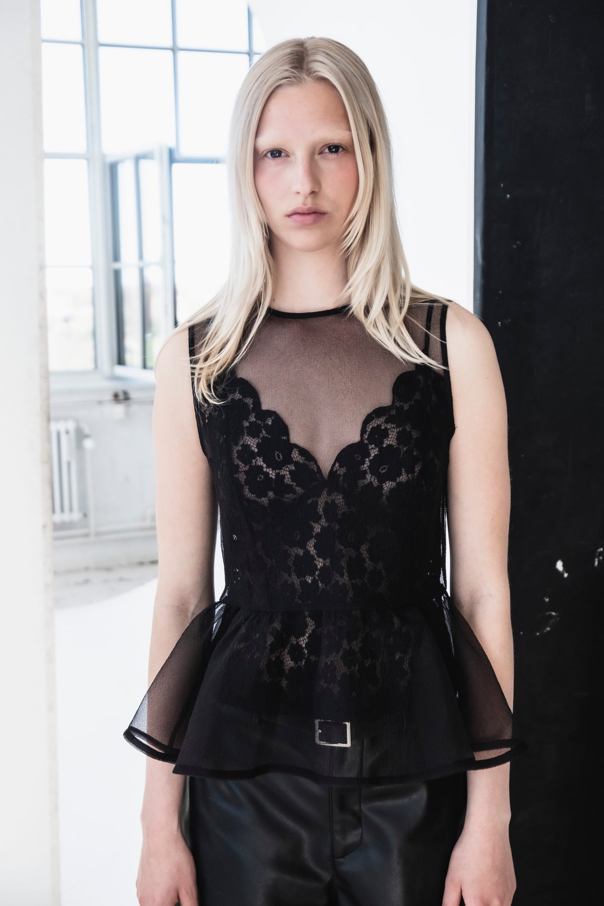 Close-up campaign portrait of a model wearing a sheer taupe-and-black top with floral lace panels, short sleeves, and a belted black skirt.