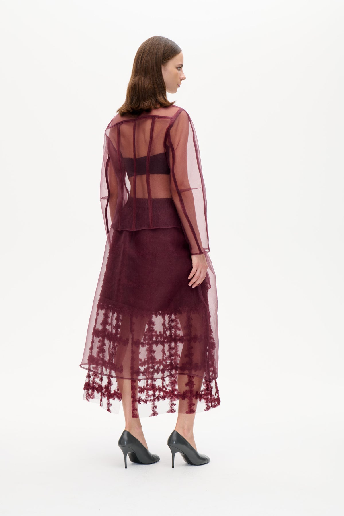 Model viewed from behind in a sheer plum long-sleeved top with panel seams, layered over a dark top and floral sheer skirt.