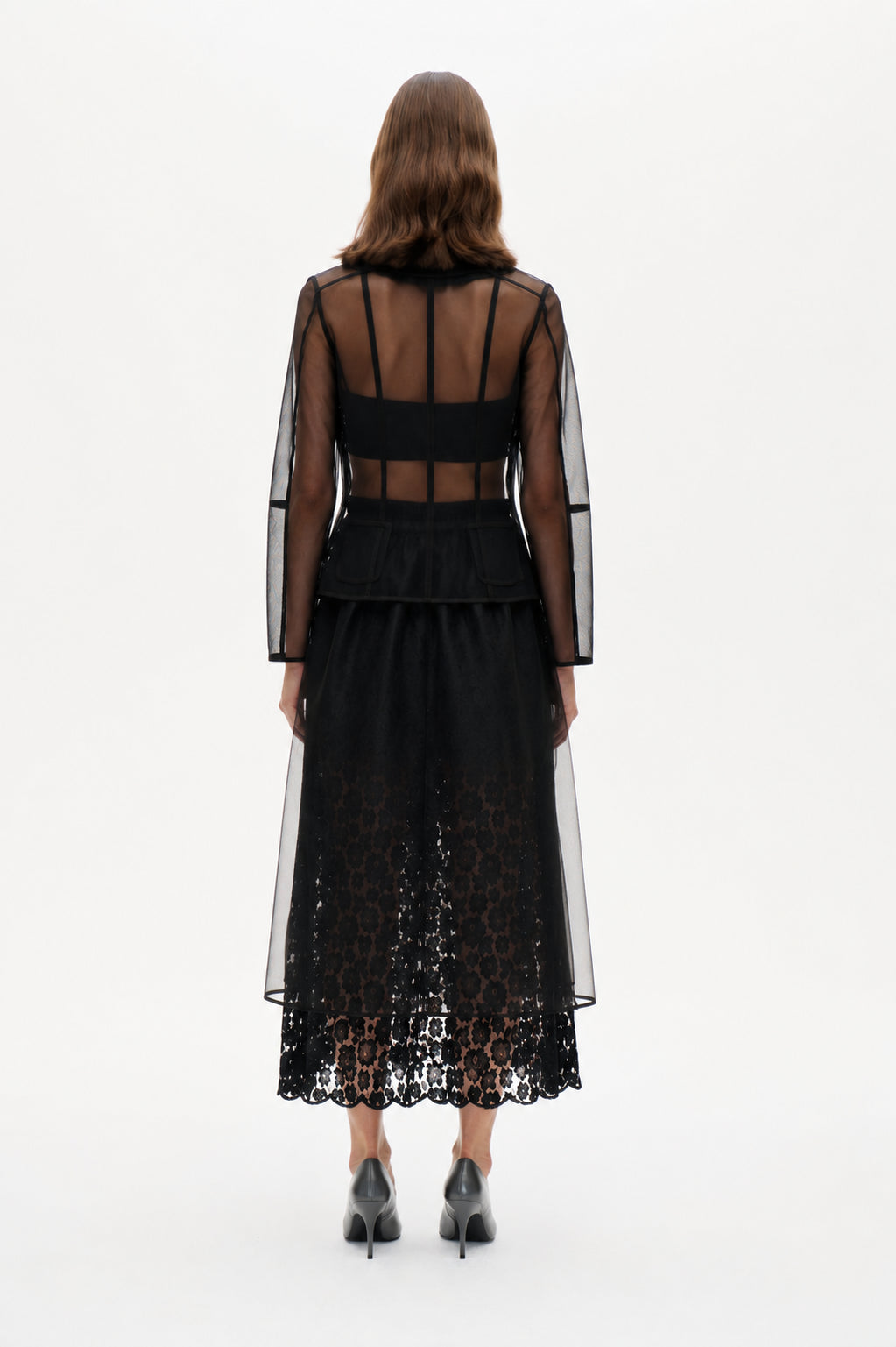 Back view of a sheer black long-sleeved jacket with panelled seams and flap pockets, layered over a black lace midi dress.