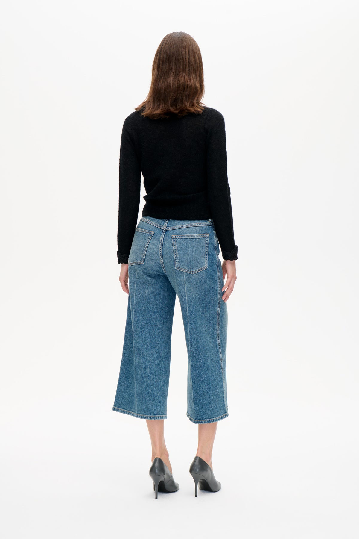 Back view of a model wearing a black long-sleeved sweater with lace cuffs and cropped wide-leg blue jeans.