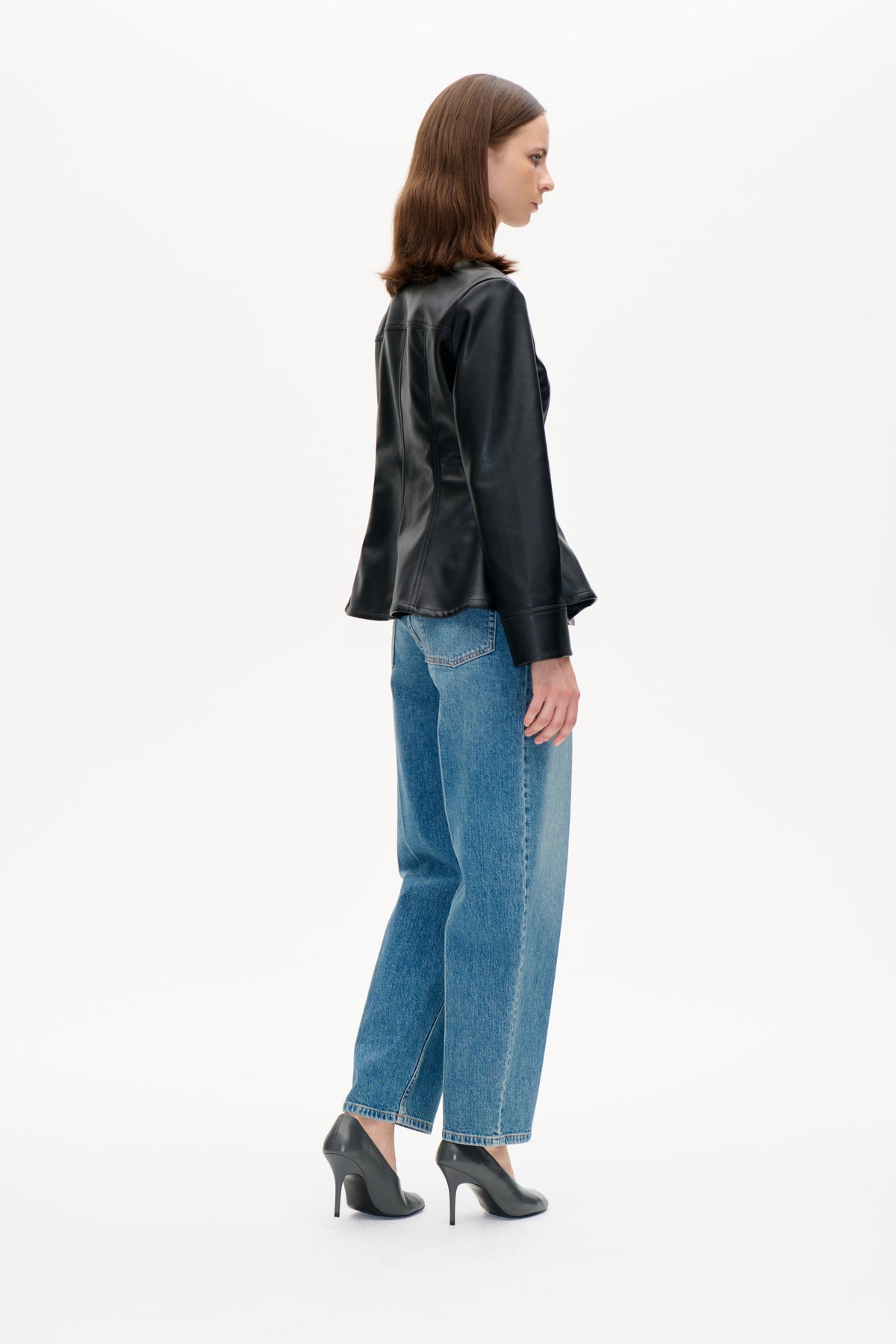 Modell im Seitenprofil trägt eine glänzende schwarze Jacke mit braunem, strukturiertem Kragen, blaue Jeans und schwarze High Heels.
