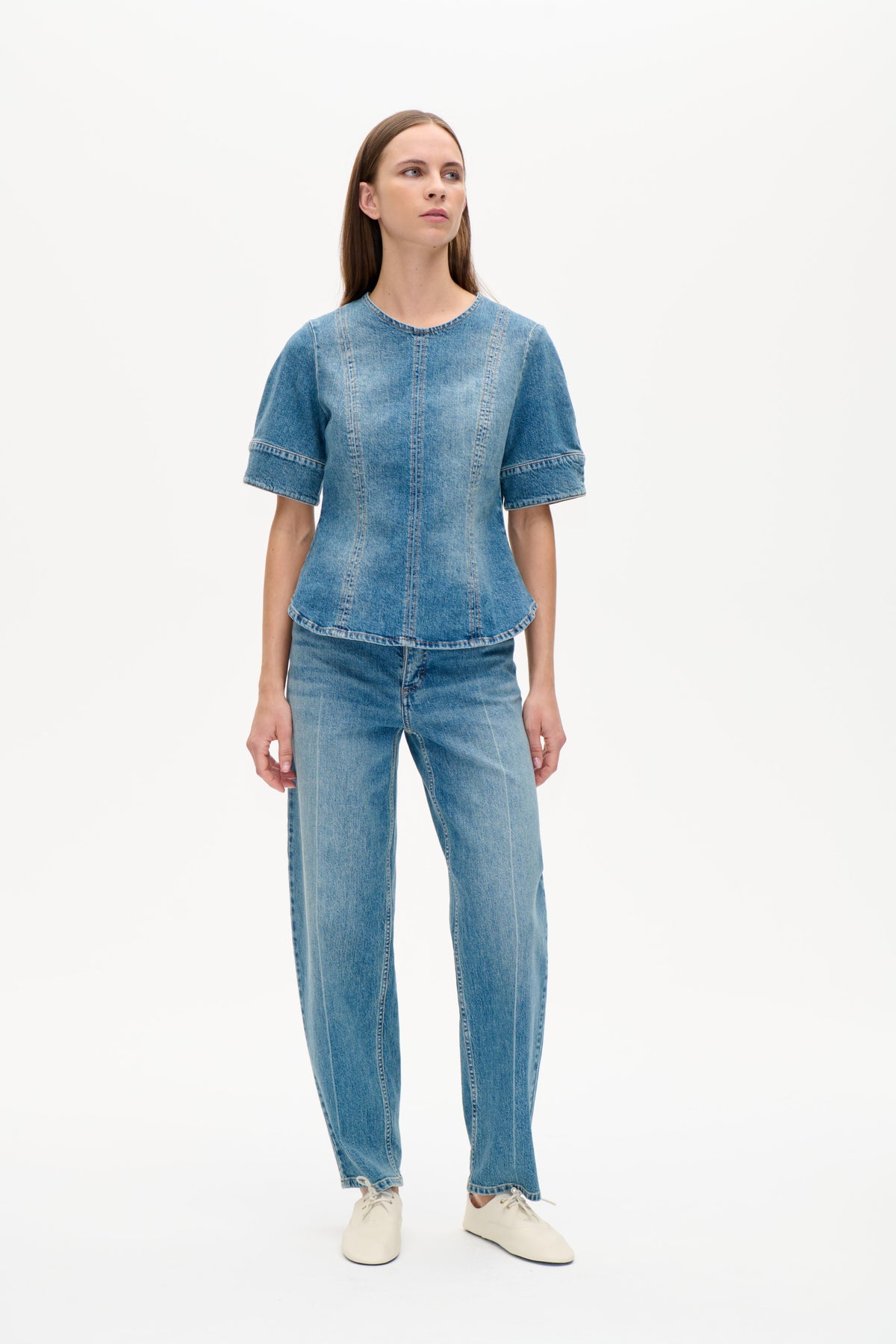 Front view of a model wearing a blue denim short-sleeve top with a round neckline and prominent vertical panel seams, paired with jeans.