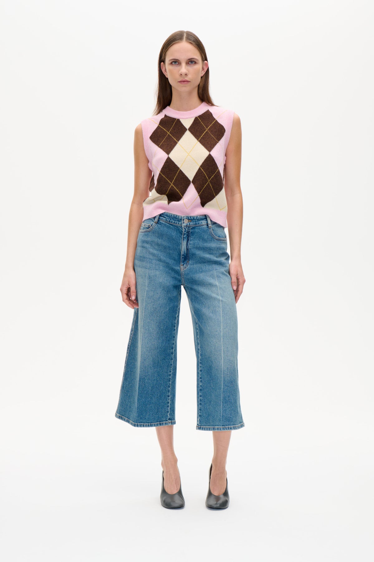 Front view of a model wearing a pink, cream and brown argyle sleeveless knit vest with blue wide-leg jeans.