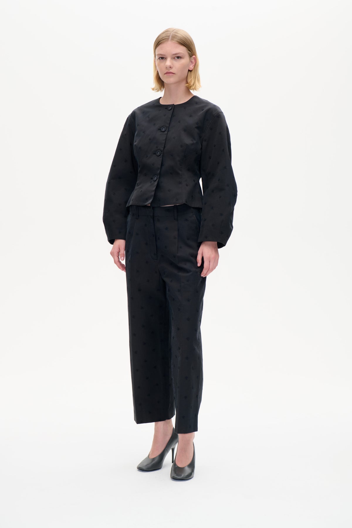 Model wearing a charcoal-black jacket with small embroidered motifs and an asymmetrical button front, paired with matching cropped trousers.