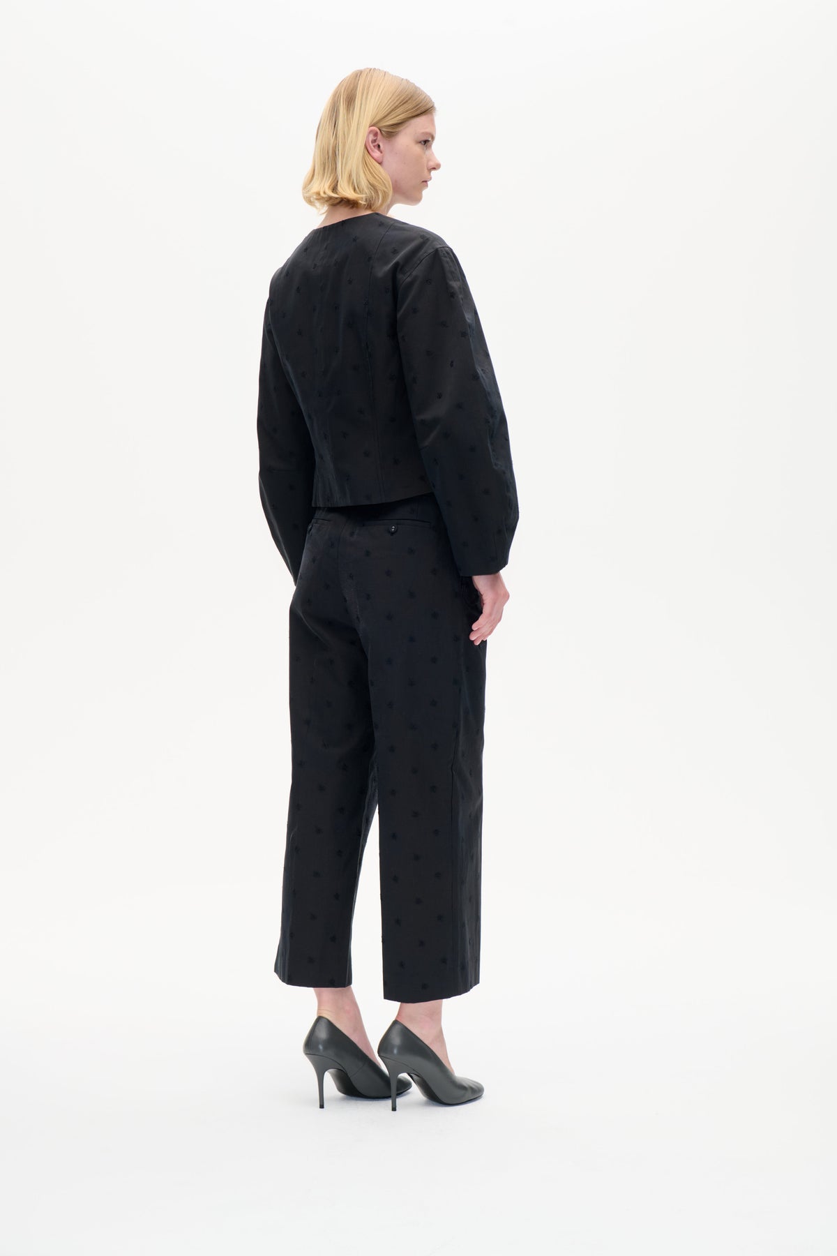Model in driekwart achteraanzicht draagt een zwarte top met lange mouwen en een cropped wijde broek met subtiele tonale motieven op een witte achtergrond.