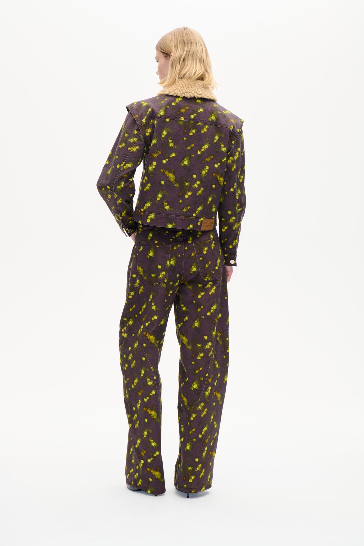 Back view of a model wearing a dark plum jacket with bright yellow-green floral print and a beige textured collar, paired with matching trousers.