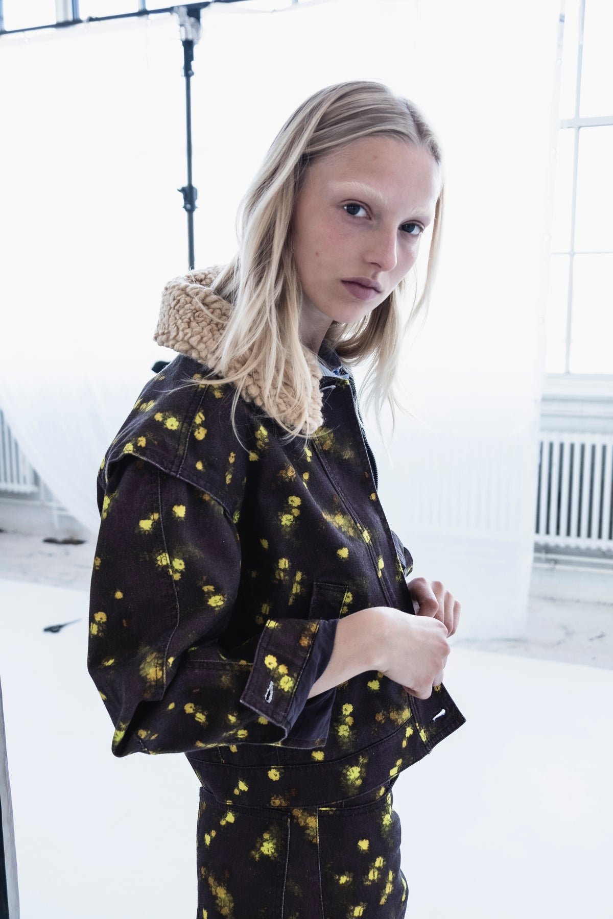 Close-up campaign portrait of a model in a dark jacket with scattered yellow floral motifs and a chunky beige knitted scarf.