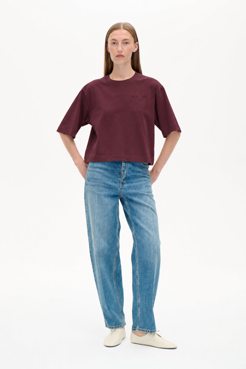 Front view of a model wearing a burgundy short-sleeve T-shirt with a tonal chest logo and light blue patterned wide-leg jeans.