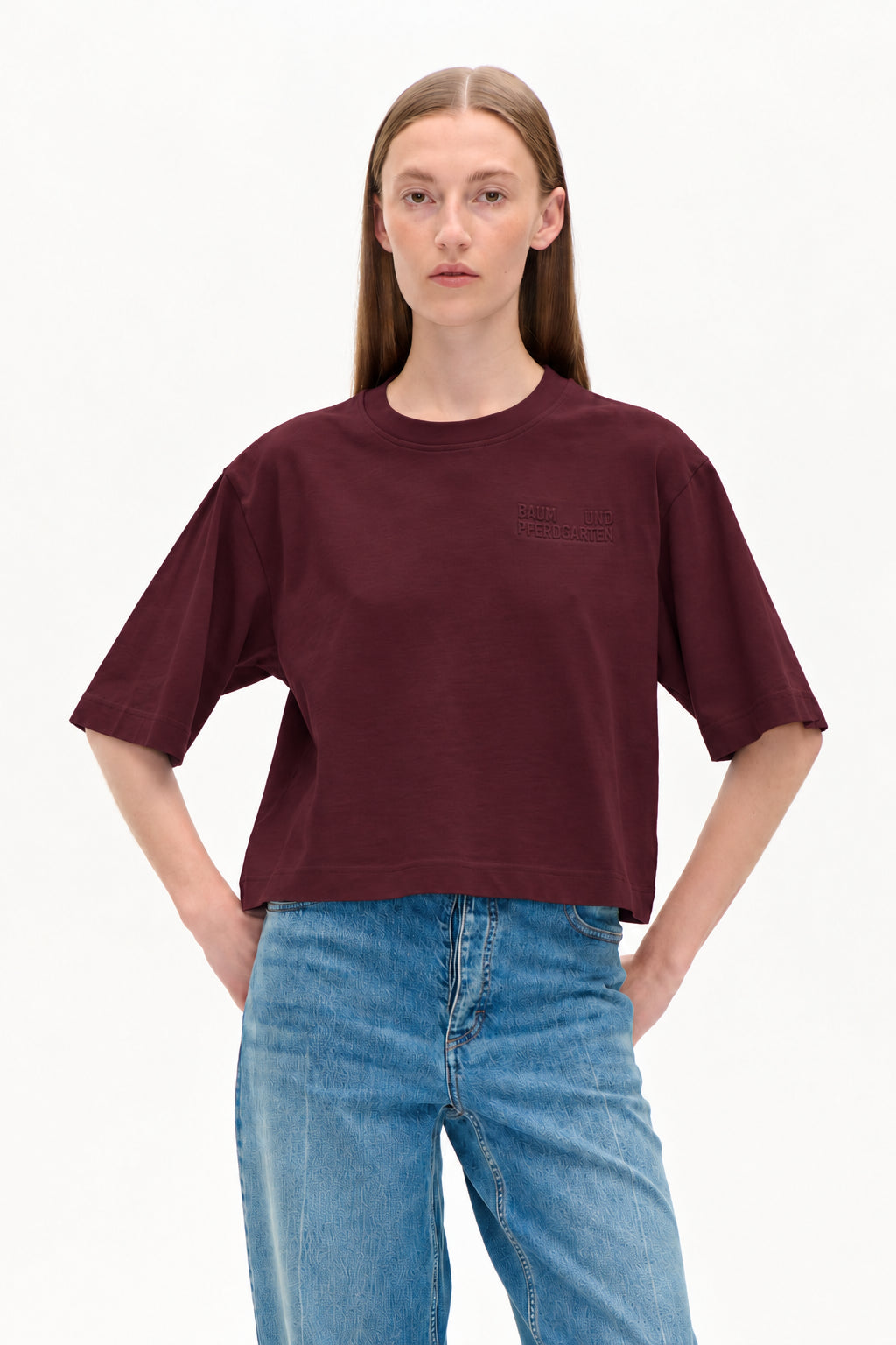 Model wearing a cropped burgundy short-sleeve T-shirt with an embossed chest logo and light blue patterned jeans.