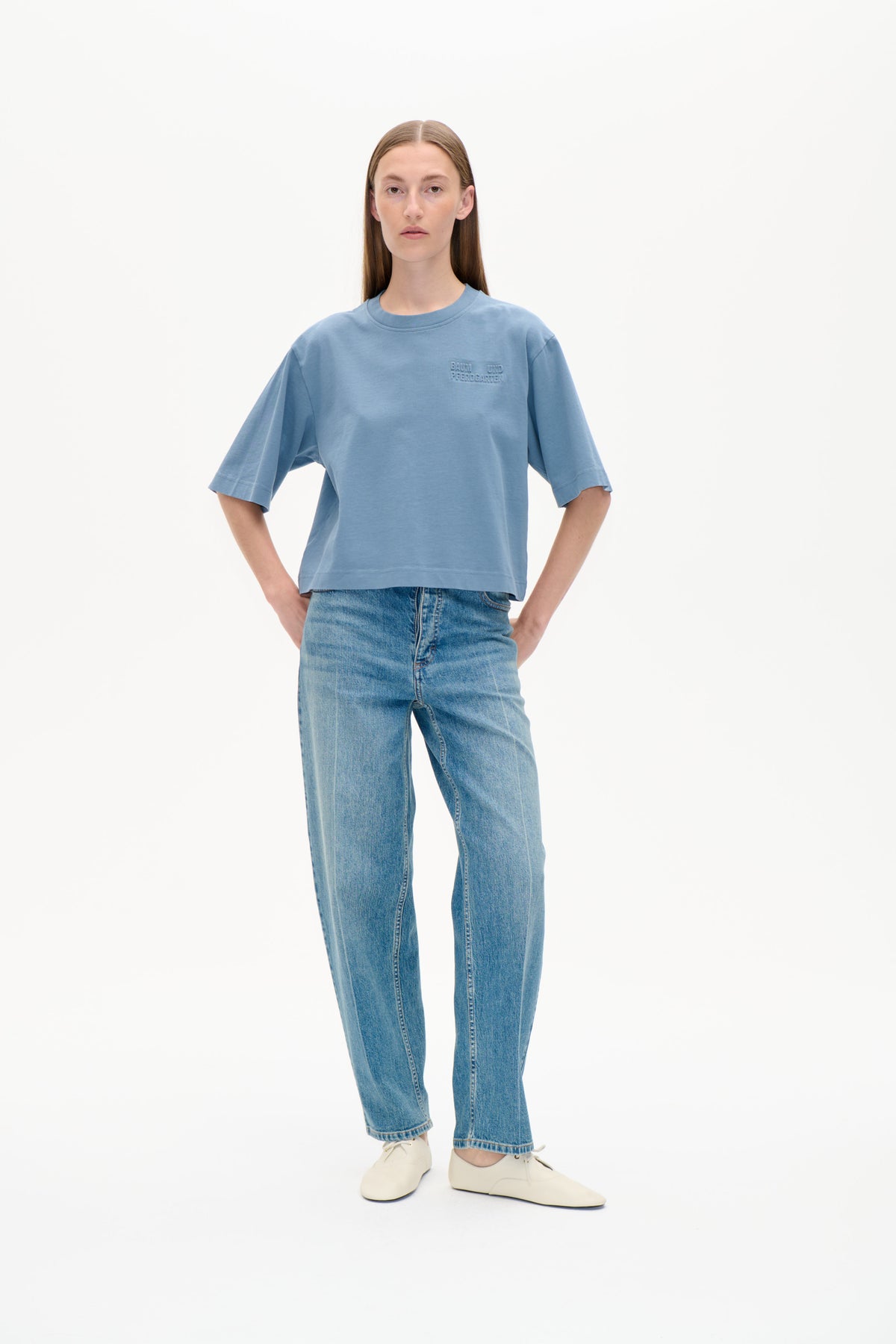Front view of a model wearing an oversized blue crew-neck T-shirt with elbow-length sleeves and a tonal chest logo, paired with jeans.