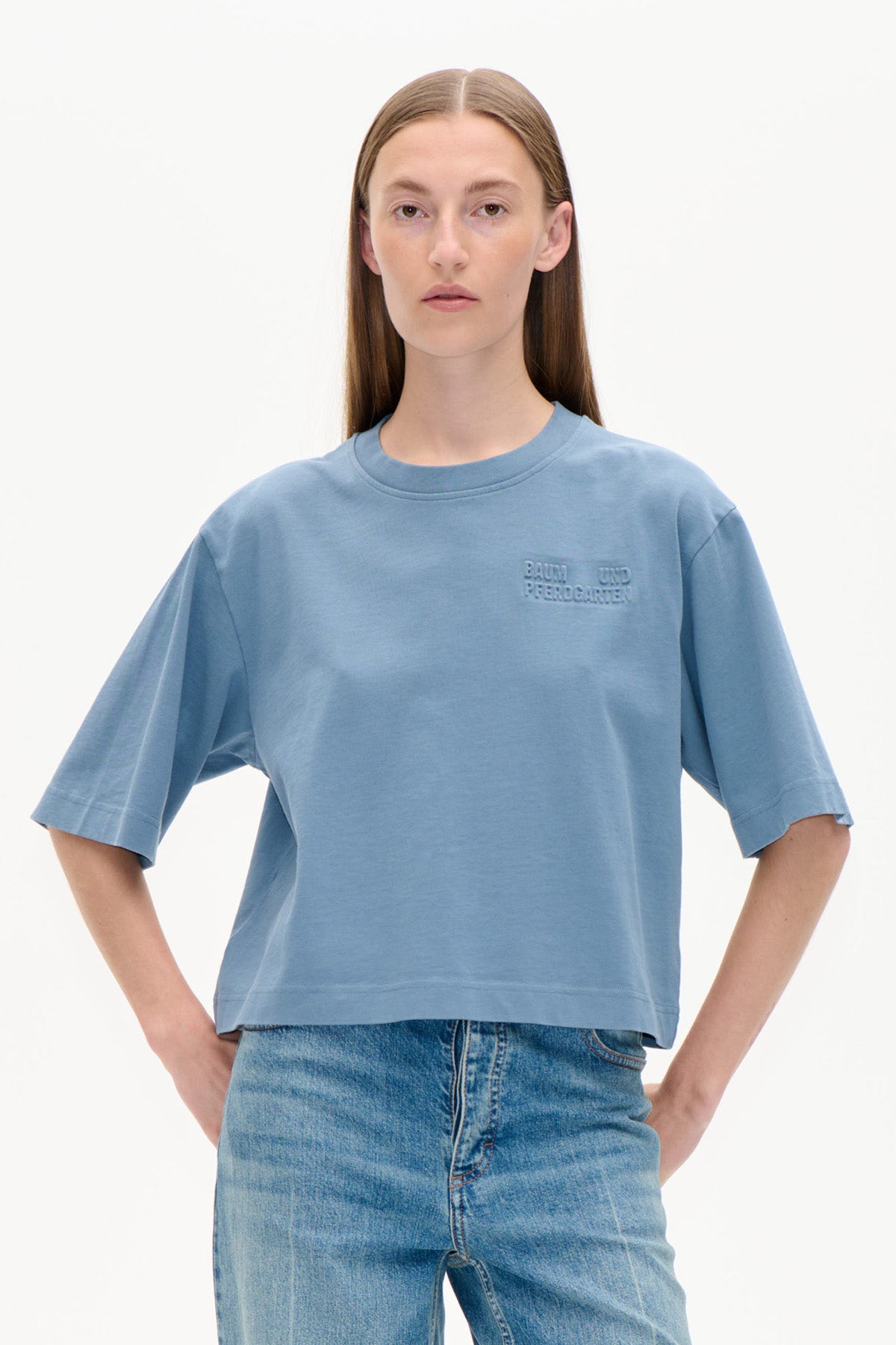 Front view of a model wearing a light blue oversized short-sleeve T-shirt with an embossed chest logo and blue jeans.