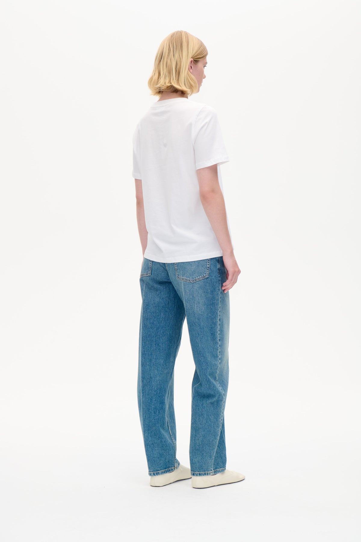 Rear view of a model wearing a plain white short-sleeved T-shirt with blue jeans and white shoes against a white backdrop.