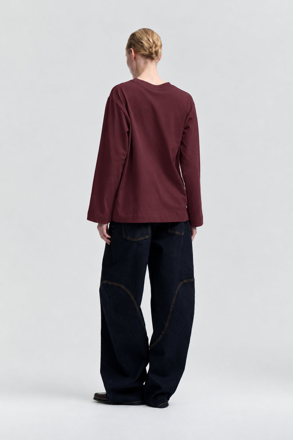 Back view of a model wearing a plain burgundy long-sleeved crew-neck top with dark wide-leg jeans.
