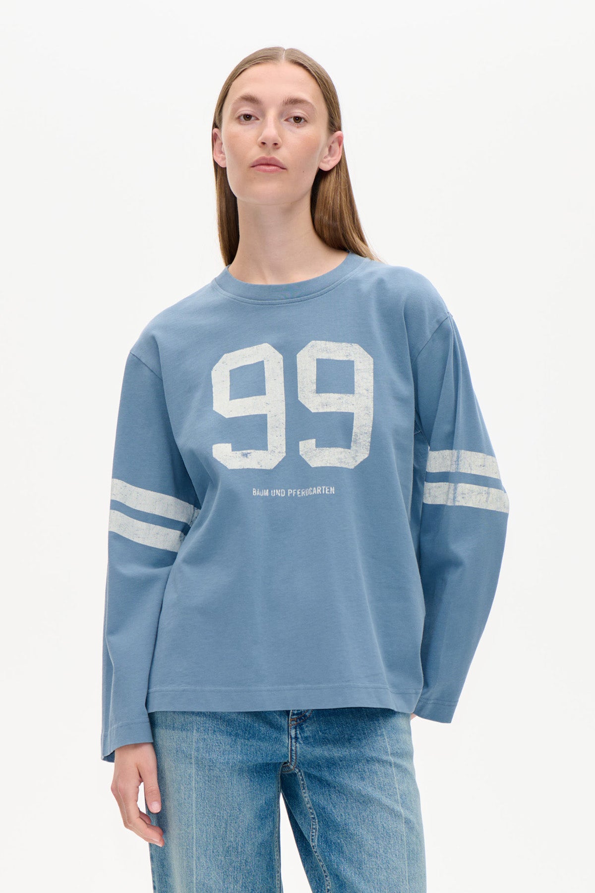 Model wearing a dusty blue long-sleeved sweatshirt with white “99” chest graphic and twin sleeve stripes, paired with jeans.