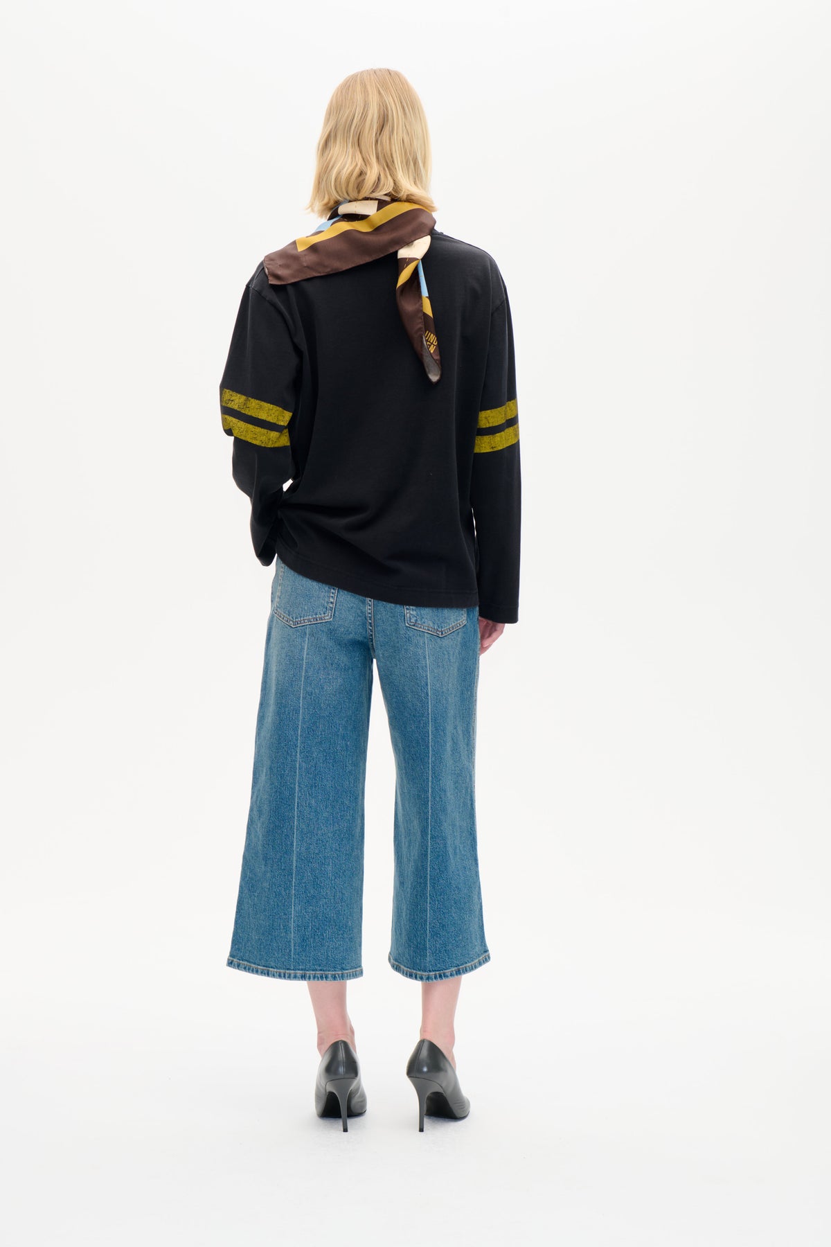 Back view of a model wearing a black long-sleeved top with mustard sleeve stripes, a multicolour geometric scarf and wide-leg blue jeans.