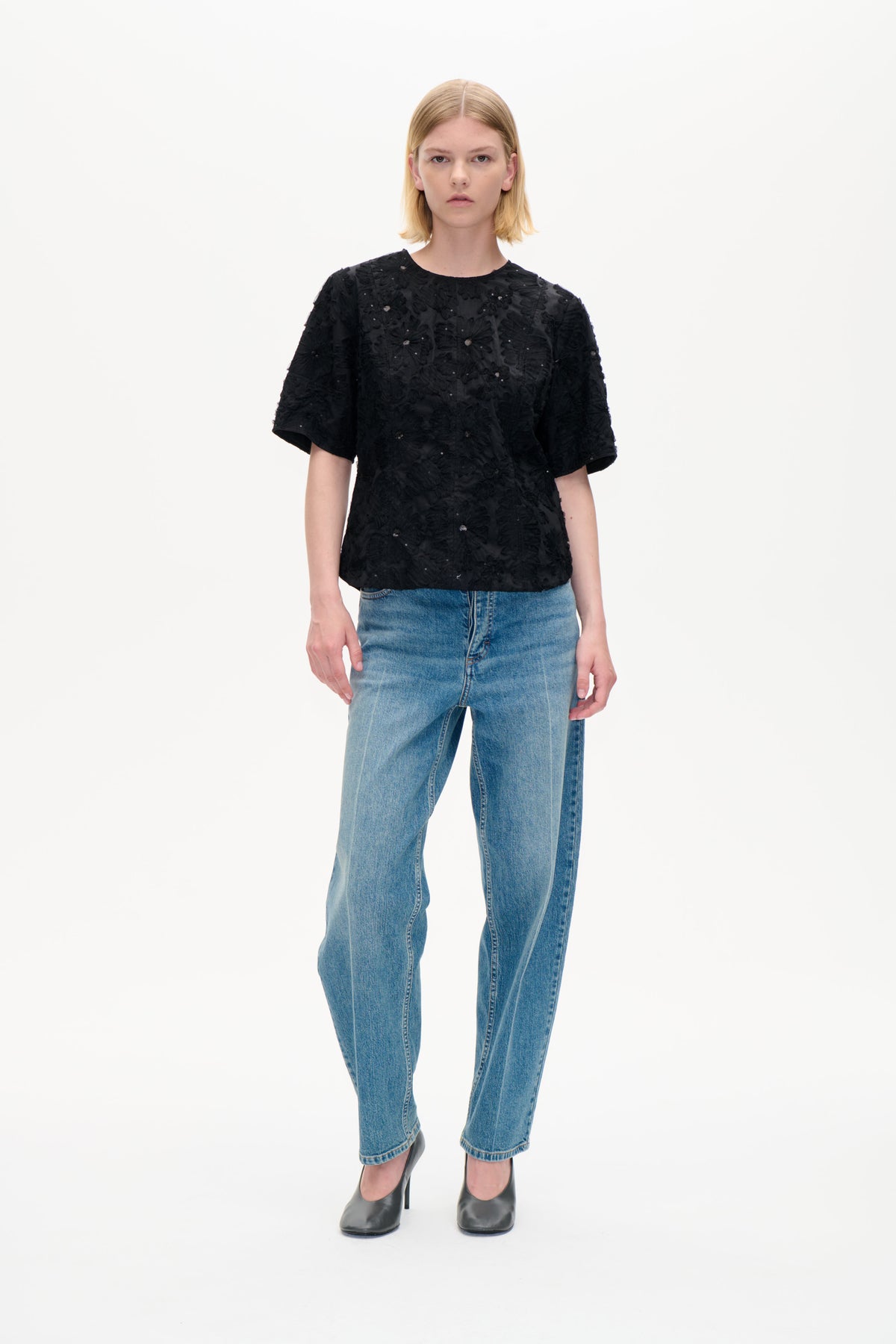 Front view of a model wearing a black short-sleeved top with raised floral detailing and small embellishments, paired with blue jeans.