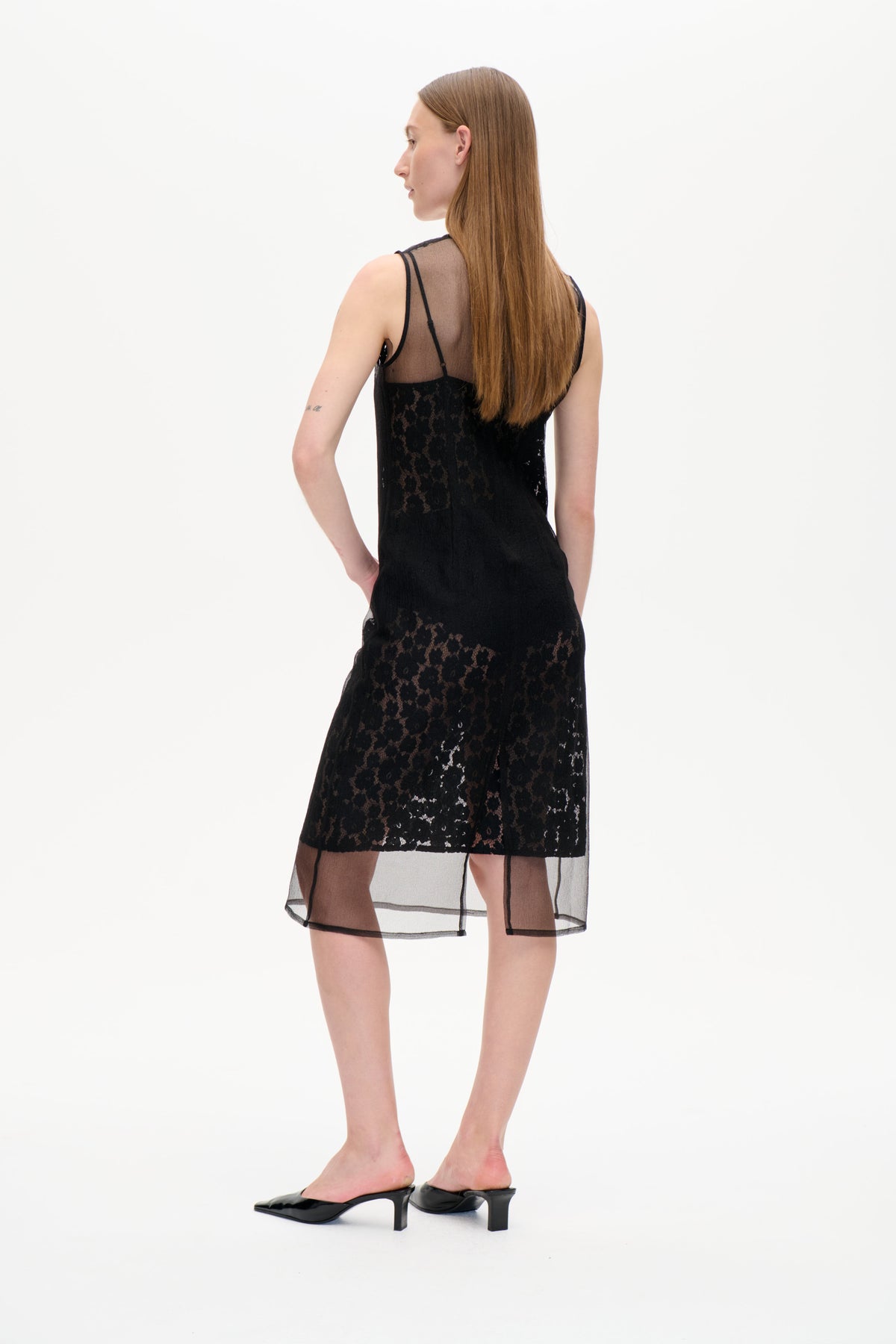 Back view of a model wearing a black lace slip dress with sheer mesh panels and a transparent, black-trimmed hem.