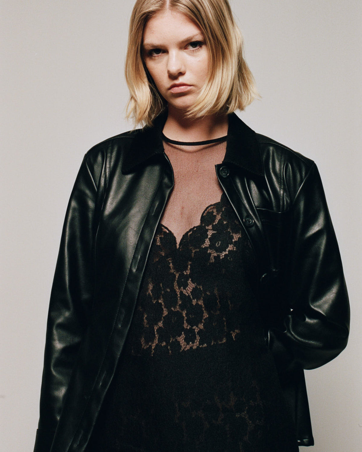 Campaign portrait of a model wearing a glossy black button-front jacket over a black floral lace dress with a sheer blush neckline.