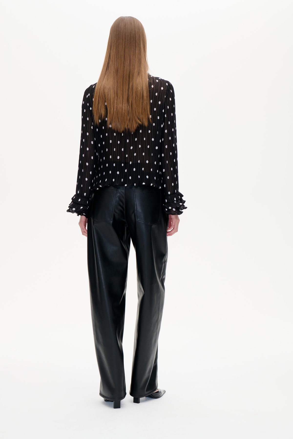 Back view of a black sheer, finely pleated blouse with white polka dots and ruffled cuffs, styled with black trousers.
