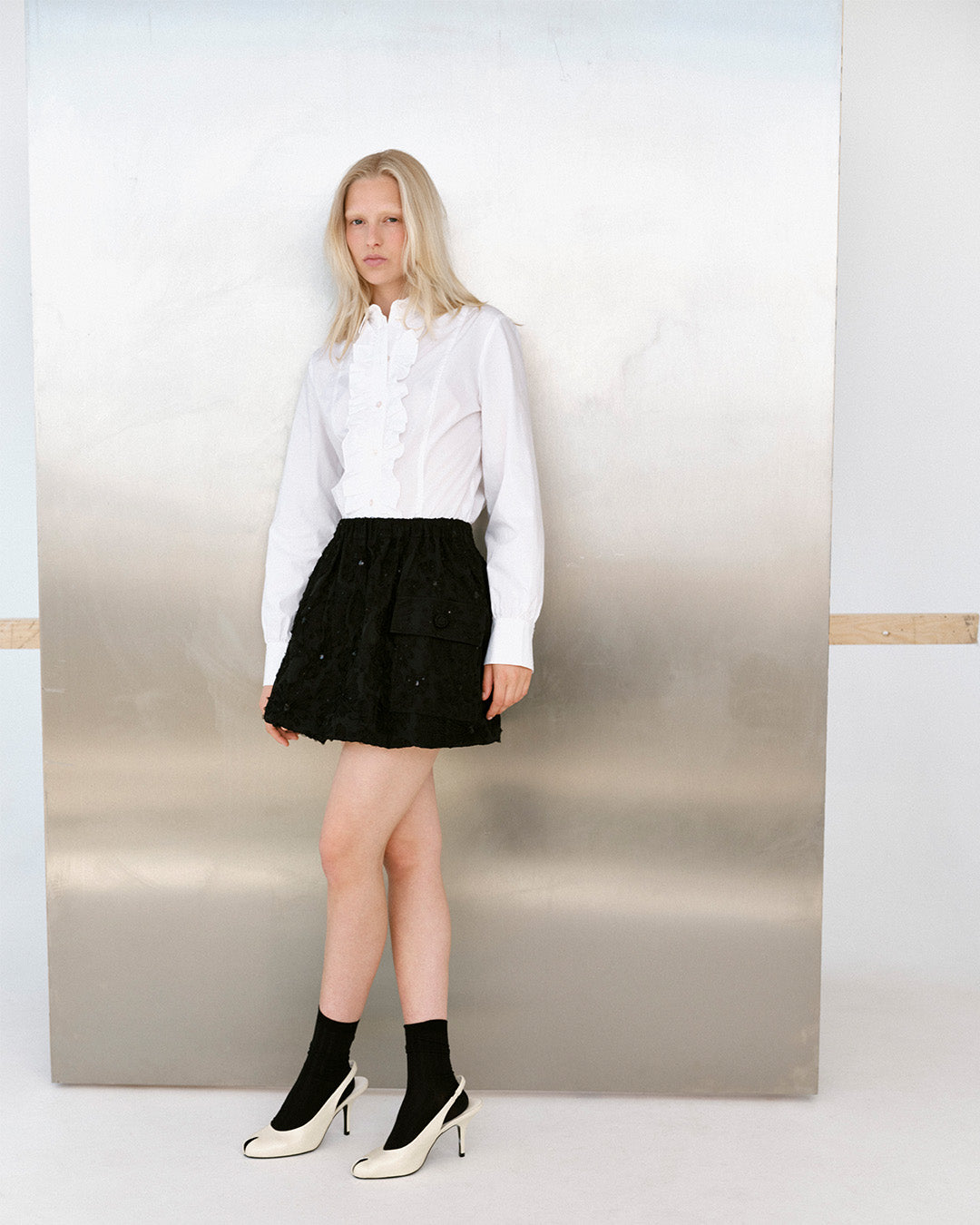 Model standing against a neutral backdrop in a white ruffle-front blouse, black textured mini skirt, black socks and white heels.